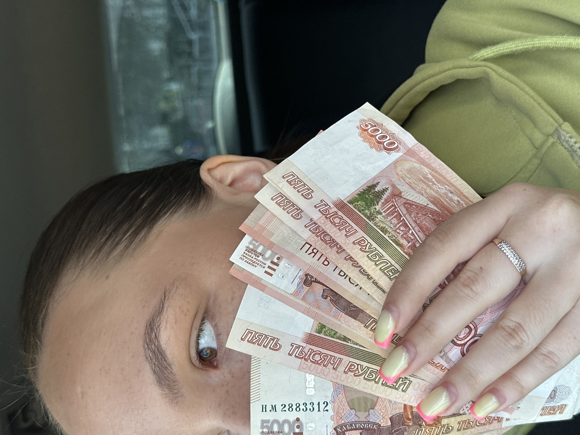 A close-up, chest-up shot shows a person's face from the nose up, peeking out from behind a fan of Russian 5,000 ruble banknotes. The person has brown hair pulled back and brown eyes. They are wearing a light green hooded sweatshirt. The banknotes are fanned out, obscuring the lower half of their face and chest. The foreground is dominated by the money and the person's hand, adorned with French manicured nails with yellow and pink tips and a diamond-studded ring on the ring finger. The background is blurred, showing dark interior of a vehicle and hints of an urban environment outside. The image appears to have been taken in Chaikovsky, Russia, as suggested by the presence of Russian currency and the general visual context.