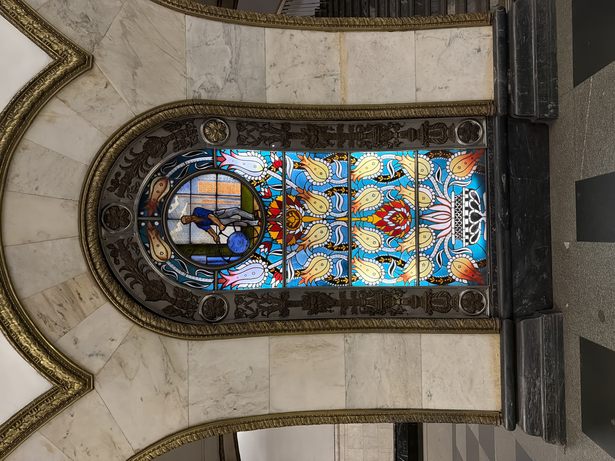 Here is a description of the image:
The image showcases a stained glass window, likely part of an architectural feature inside a building, possibly a subway station in Moscow, Russia. The window is set within an ornate framework of marble and metal, featuring decorative carvings and details.
The stained glass itself is divided into sections. One portion depicts a figurative scene of a worker, while the other parts features patterns of stylized tulips in a variety of colors set against a blue background.
The surrounding architectural elements are heavily decorated with gold trim and marble cladding. The image is captured at a slightly tilted angle.