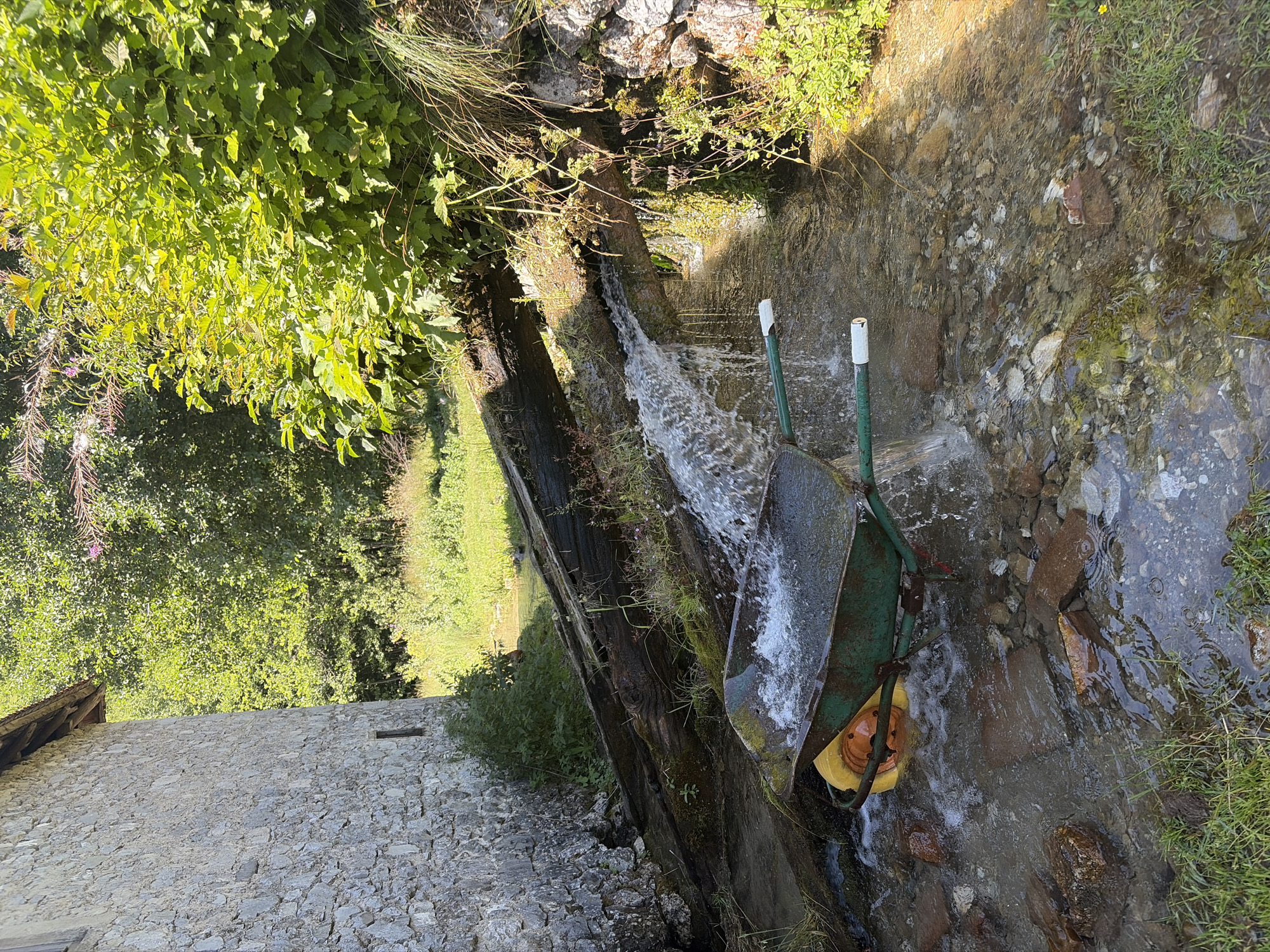 This image depicts a scene in Gusinje, Montenegro, featuring an old stone building on the left and lush green foliage on the right. In the foreground, a green wheelbarrow filled with water is positioned in a shallow stream flowing over rocks and pebbles. A large, weathered log spans the stream, serving as a makeshift bridge or channel for the water. The water cascades over the log, creating a waterfall effect into the stream. The overall environment appears natural and rustic, suggesting a rural or historical setting.