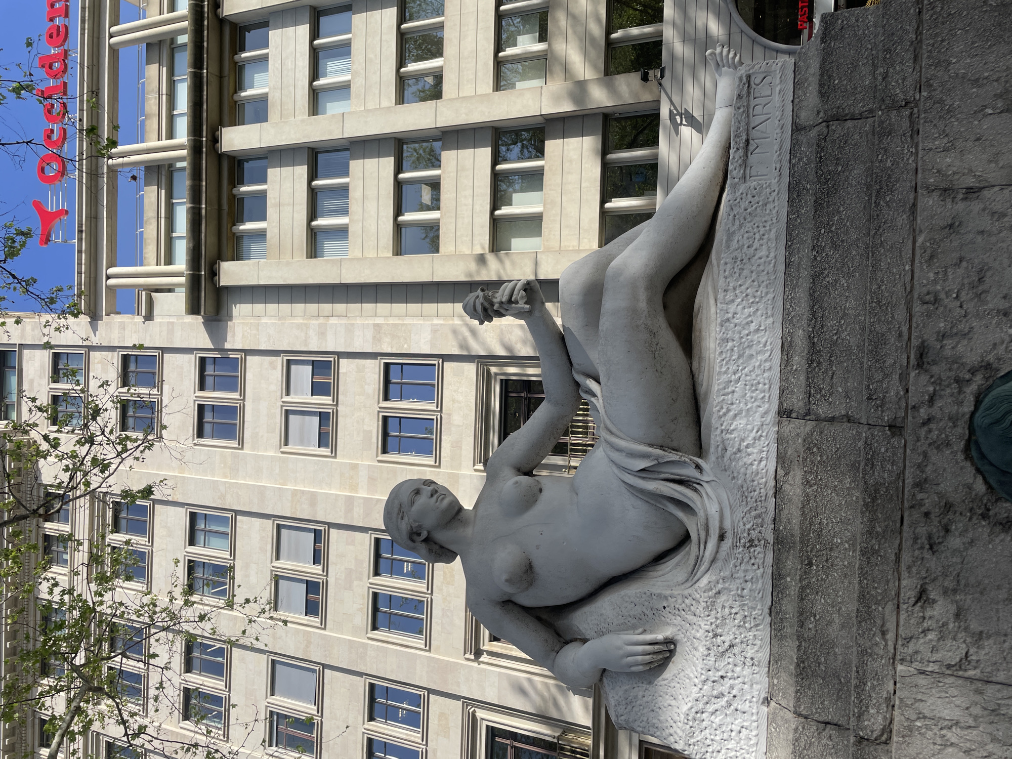 The image shows a reclining nude statue of a woman, possibly made of marble or a similar light-colored stone. The statue is placed on a stone base with inscriptions on it. The background features a tall building with a grid-like facade of windows, some showing the reflection of the blue sky. A partial view of a tree on the left side suggests the presence of greenery nearby. Part of a red sign with the word "Occidenten" is visible at the top left of the image.
Based on the architectural style and sign, this appears to be the sculpture "F. Mares" located in Barcelona, Spain.