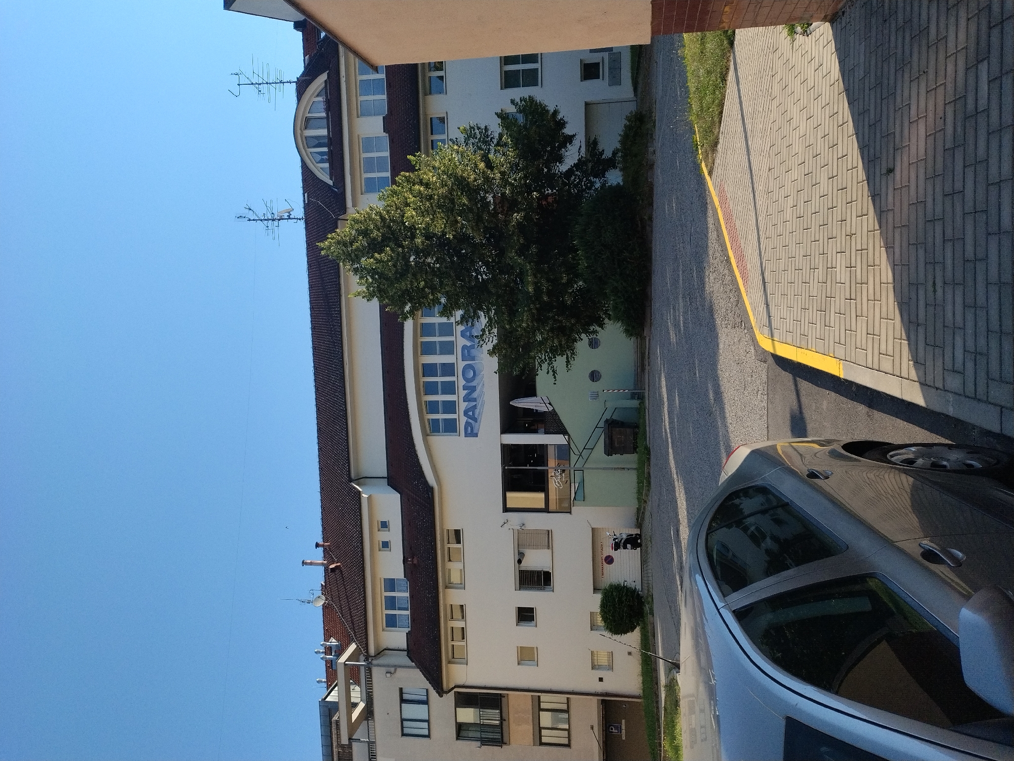 This image depicts a clear, sunny day in Hluboká nad Vltavou, Czechia. In the foreground, a silver sedan is parked on a paved driveway. The car is angled towards the right of the frame, with its driver's side door and windows clearly visible. Beyond the car, a building with a white facade dominates the scene. The building has multiple floors and a brown tiled roof. A large green tree stands in front of the building, partially obscuring a section of its facade. Above the entrance to the building, the word "PANORAMA" is displayed in large blue letters. A set of stairs leads up to the entrance, which appears to be a business with a window displaying the word "Bistro." To the left of the building, a smaller, multi-story structure with a beige facade can be seen. The background is a clear blue sky. The environment is urban, with paved surfaces and some greenery. There are no people visible in the image.