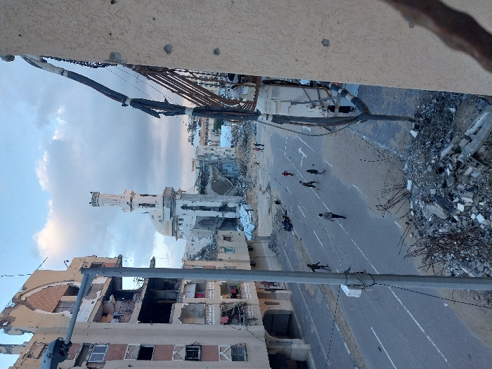 An elevated view captures a street scene in Khan Yunis, Palestinian Territory, depicting extensive structural damage to the surrounding urban environment. In the center-background stands a large mosque with a prominent minaret and dome, exhibiting severe damage with sections of its facade and structure missing, and debris accumulated around its base. To the left, a multi-story residential building shows significant destruction, with collapsed walls, exposed interiors, and shattered windows across multiple floors; an arched architectural detail is visible on its upper section.
The street itself is paved with intact white lane markings. Approximately ten individuals are present on the road: four figures, appearing to be children or adolescents, are actively engaged in playing soccer with a light-colored ball in the middle of the street. Two individuals are near a parked motorcycle on the left side of the road, while others are seen walking or standing.
The immediate foreground on the right features a portion of a heavily damaged building wall with visible rebar and multiple circular impact marks, consistent with bullet or shrapnel damage. Above this, numerous utility cables are dislodged and hanging loosely. Piles of concrete rubble, twisted metal, and other building debris line the sides of the street, particularly in the foreground. A gray street lamp post with attached wiring stands on the left side of the street. The sky above is partially cloudy.