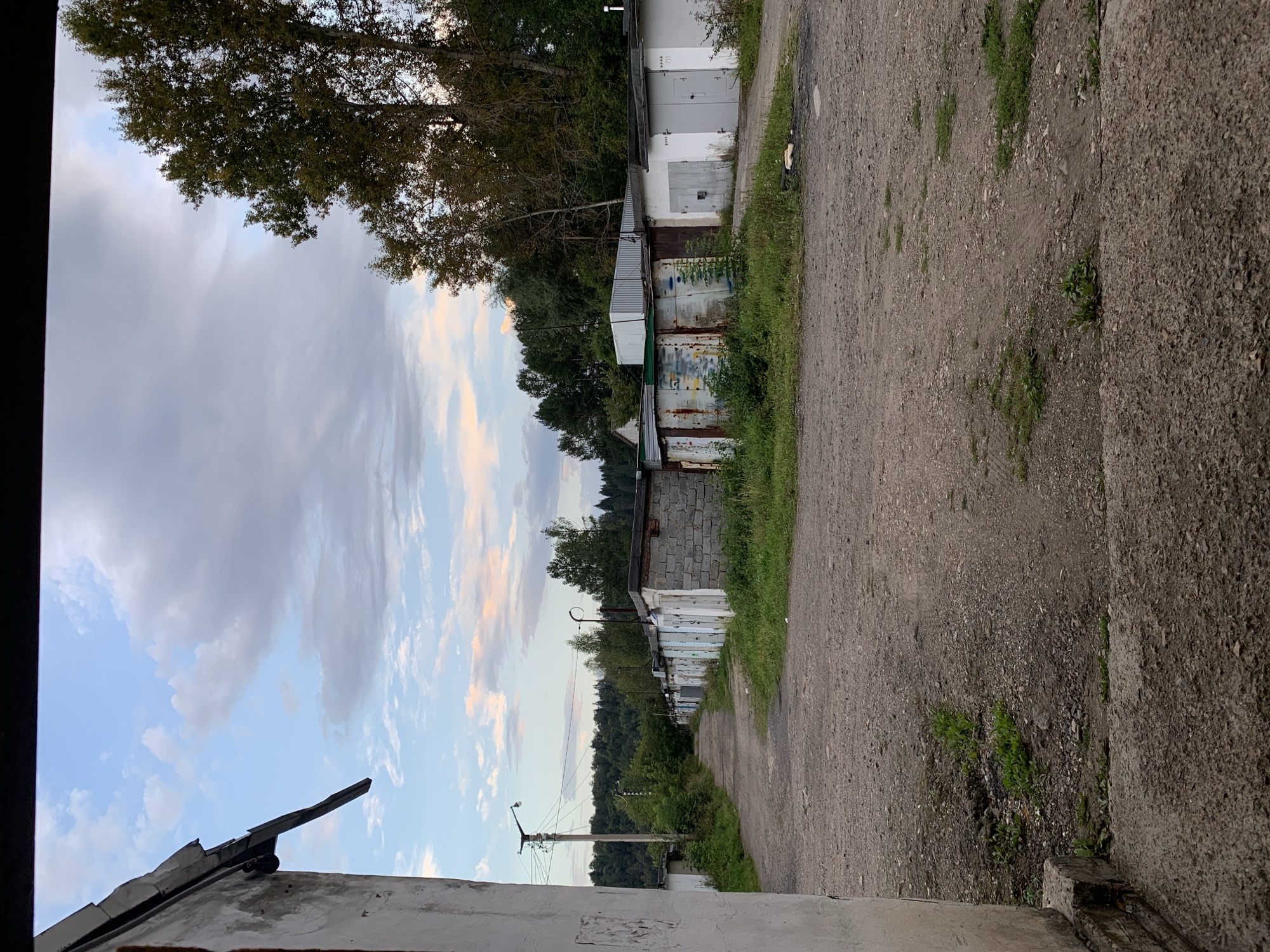 An overcast sky with pastel clouds is visible above a row of garages and a dirt road in the city of Asha, Russia. The garages appear to be made of brick and metal. Some are weathered and rusted, while others are painted white. Vegetation, including grass and trees, grows around the garages and along the sides of the road. In the distance, a dense forest covers rolling hills. The foreground shows a portion of a concrete structure, possibly a foundation or sidewalk. No people or vehicles are present in the image.