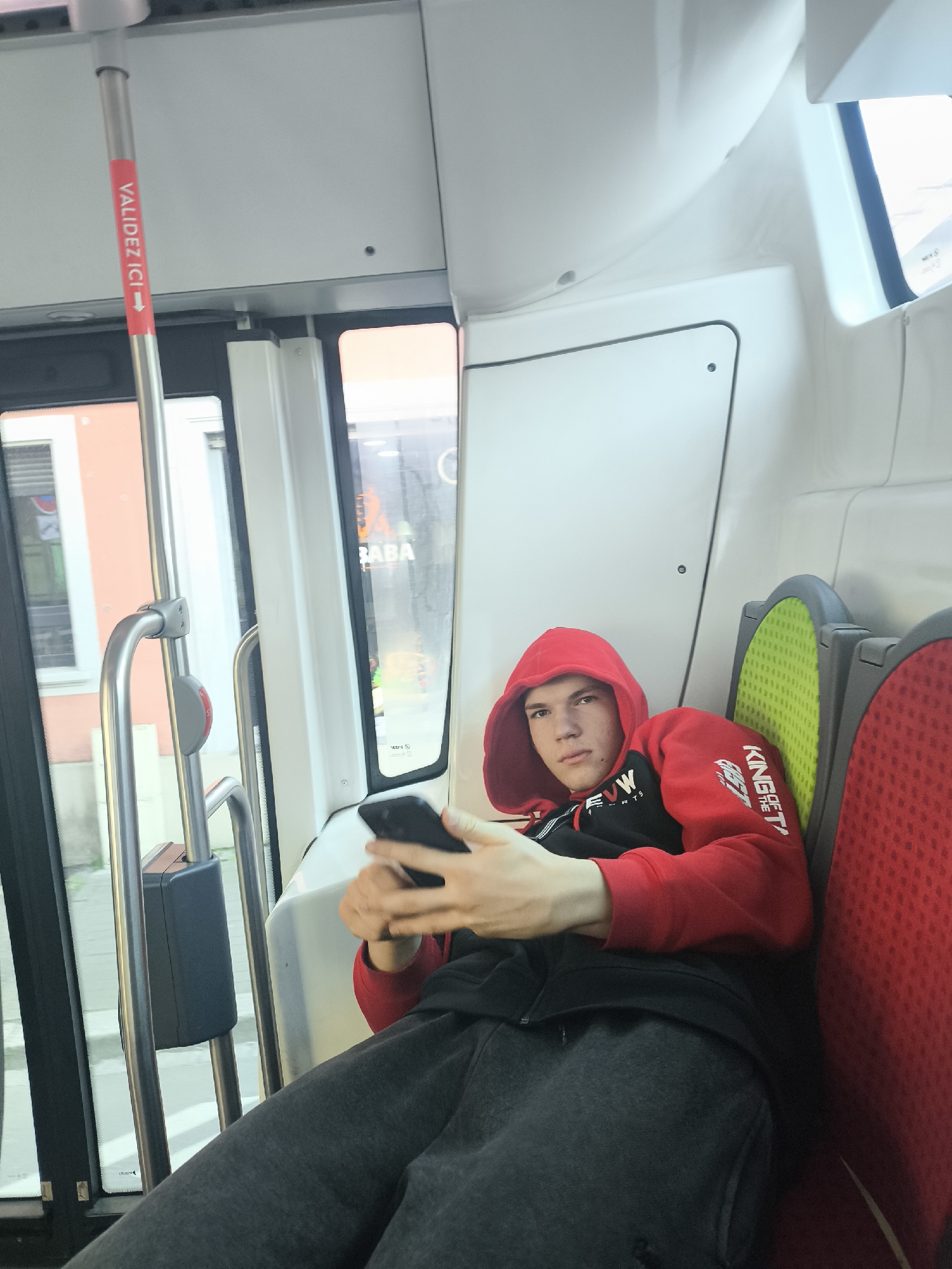 A young man is seated inside what appears to be a public transport vehicle, possibly a tram or bus. He is wearing a red hooded sweatshirt over a black long-sleeved shirt and dark gray sweatpants. His hood is up. He is looking directly at the camera with a neutral expression, holding a black smartphone in both hands. He is leaning against the back of a seat.
The interior of the vehicle is bright, predominantly white and light gray, with visible windows to the left showing an exterior urban or suburban scene. Through the window, parts of buildings with a light orange or pink facade and other structures are visible. There's a sign outside that partially reads "BABA".
To the right of the man are two seats, one with a lime green textured backrest and the other, partially visible, with a red textured backrest. Metal poles and grab handles typical of public transport are prominent on the left side of the frame. One vertical pole has a red and white sticker wrapped around it with an arrow and the text "VALIDEZ ICI" (Validate Here), indicating a ticket validation point.
The lighting suggests it is daytime. The overall scene depicts a casual moment of travel. On the man's red hoodie, white text is visible on the sleeves: "KING OF THE T" on the right sleeve and "EW RTS" (likely part of "NEW SPORTS" or similar) on the left sleeve.