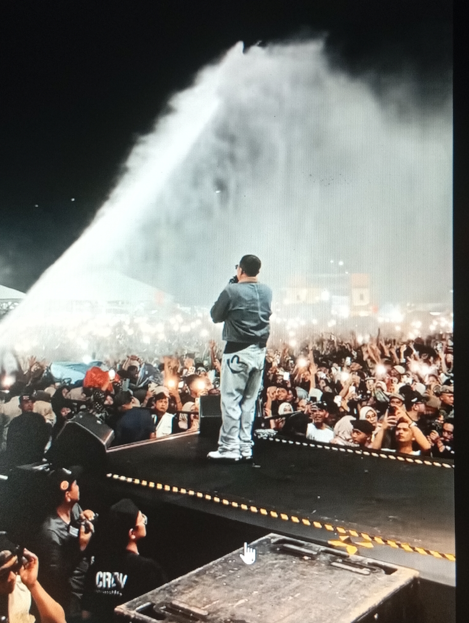 The image shows a male performer on stage at an outdoor concert, facing away from the camera and towards a large crowd. The performer is wearing a grey jacket and light-colored pants. A significant amount of mist or smoke is being emitted from a device on stage, creating a dramatic effect and partially obscuring the performer and the crowd. The crowd, composed of many people, is illuminated by numerous lights held up by audience members. Some people in the crowd are raising their hands. The stage has speakers and other equipment. The background suggests an urban environment, potentially a public space or stadium within Lhokseumawe, Indonesia, given the context. The lighting indicates it is nighttime.