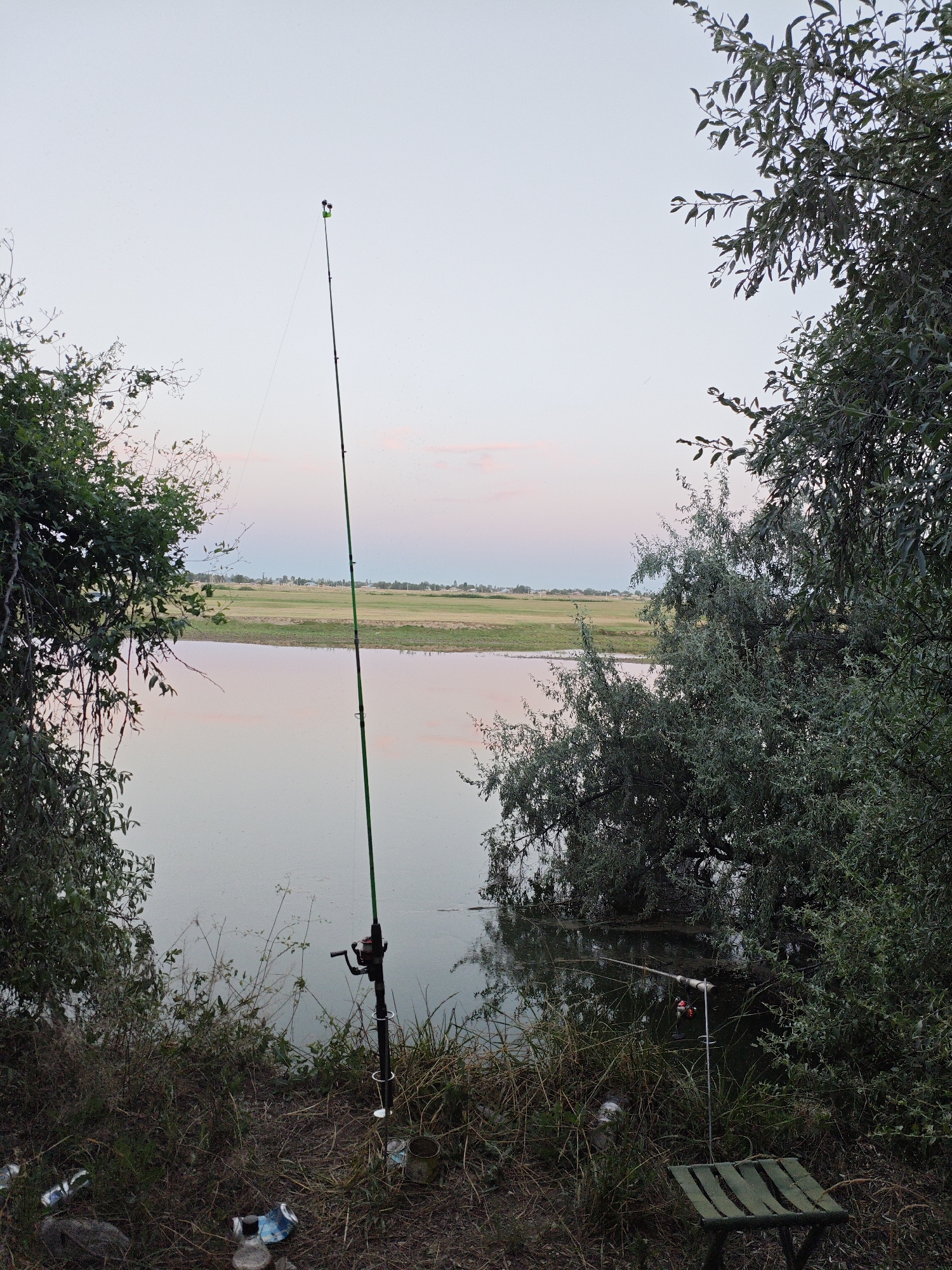 An outdoor scene depicts a body of water, possibly a river or lake, with lush green foliage on the left and right sides of the frame. A fishing rod with a reel is planted vertically in the ground near the water's edge. A small, foldable green stool is positioned to the right of the fishing rod, and several discarded cans are visible on the ground in the foreground. The sky displays a soft gradient of pink and pale blue, suggesting either sunrise or sunset. In the distance, across the water, there is a flat, green expanse of land dotted with a few buildings. The overall impression is one of a tranquil, natural setting, likely a popular fishing spot.
