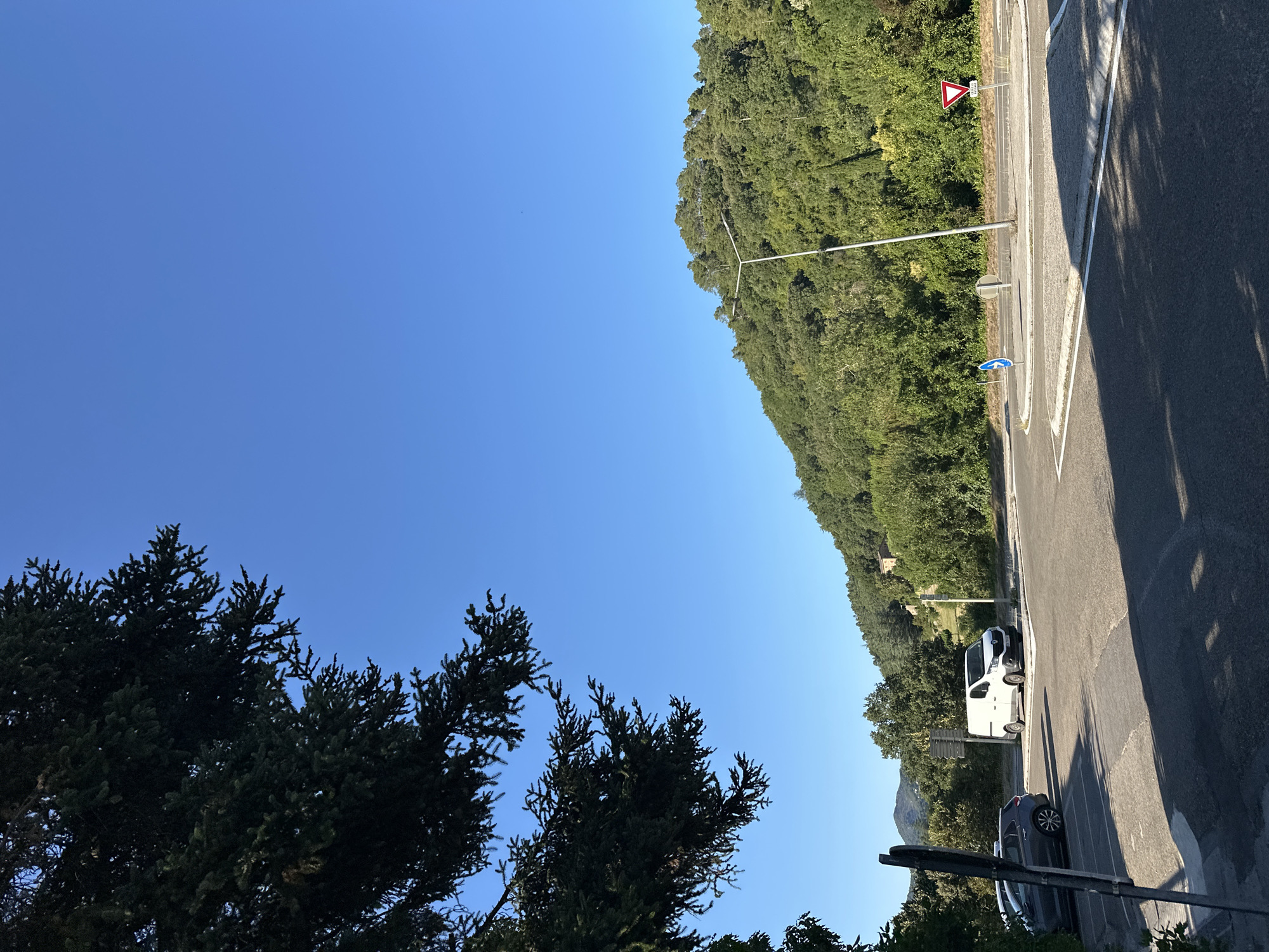 The image depicts a rural intersection in Générargues, France, under a clear blue sky. A dense, green hillside covered in trees dominates the background. In the foreground, a paved road curves to the right, forming what appears to be a roundabout. A white van is parked on the side of the road, and a dark grey car is parked to its left, partially obscured by foliage and a signpost. A modern streetlight stands in the center of the road. Traffic signs are visible: a blue circular sign with a white arrow pointing right indicates traffic flow, and a red and white triangular sign with a red border signifies a yield. The overall scene suggests a quiet, sunny day in a village setting.