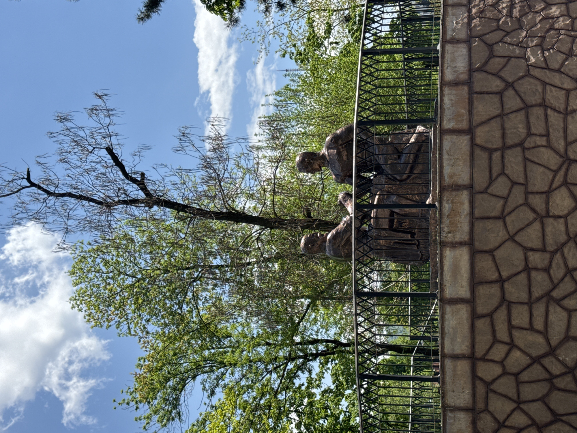 The image shows a bronze statue of two figures, seemingly looking down from a balcony, in the city of Vladikavkaz, Russia. The balcony has a decorative black metal railing. A wall made of irregularly shaped stones is adjacent to the balcony. Behind the balcony, there is foliage consisting of green trees and some bare tree branches. The sky is blue with scattered white clouds.