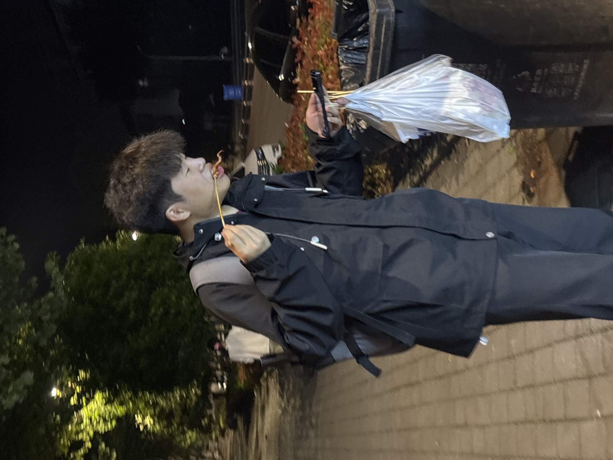 A male with dark, curly hair and a black jacket is eating food on a stick in Jiangjin, China. He holds a plastic bag and other food skewers in his left hand. The background includes a brick walkway, trees, and a dark vehicle or structure. The image is taken at night.