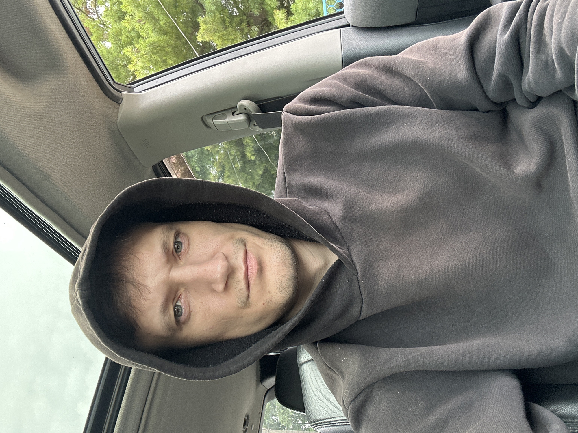 A man wearing a dark hooded sweatshirt sits in the driver's seat of a car, looking directly at the camera. He has short brown hair and light-colored eyes. The car's sunroof is open, revealing a cloudy sky. Trees are visible through the car window. The interior of the car, including the dashboard and a seatbelt, is visible. The scene appears to be outdoors in a wooded area, possibly on the outskirts of Makiv, Ukraine, given the context.