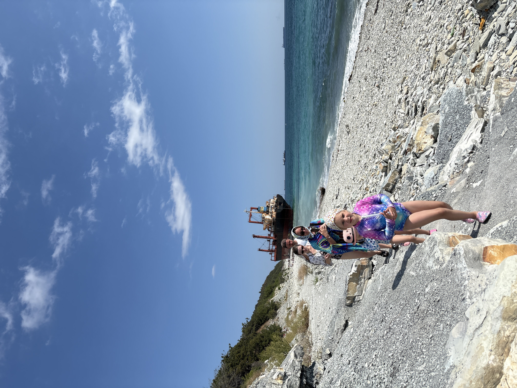 A group of four people are walking along a rocky shoreline with the sea on their right. A large cargo ship is anchored in the distance. The sky is blue with scattered white clouds. The person in the foreground is a young girl wearing a colorful swimsuit and a headscarf. She is smiling and looking at the camera. Behind her, a woman in a patterned scarf and sunglasses is holding a pink bag. Next to her, a man and another woman are walking. The shoreline is covered in small, grey and white stones, with larger rocks and some sparse vegetation on the left side of the image. In the background, other ships are visible on the horizon. The overall scene depicts a sunny day at the coast. This is likely the coast near the shipwreck of the Rio de Janeiro in Kabardinka, Russia.