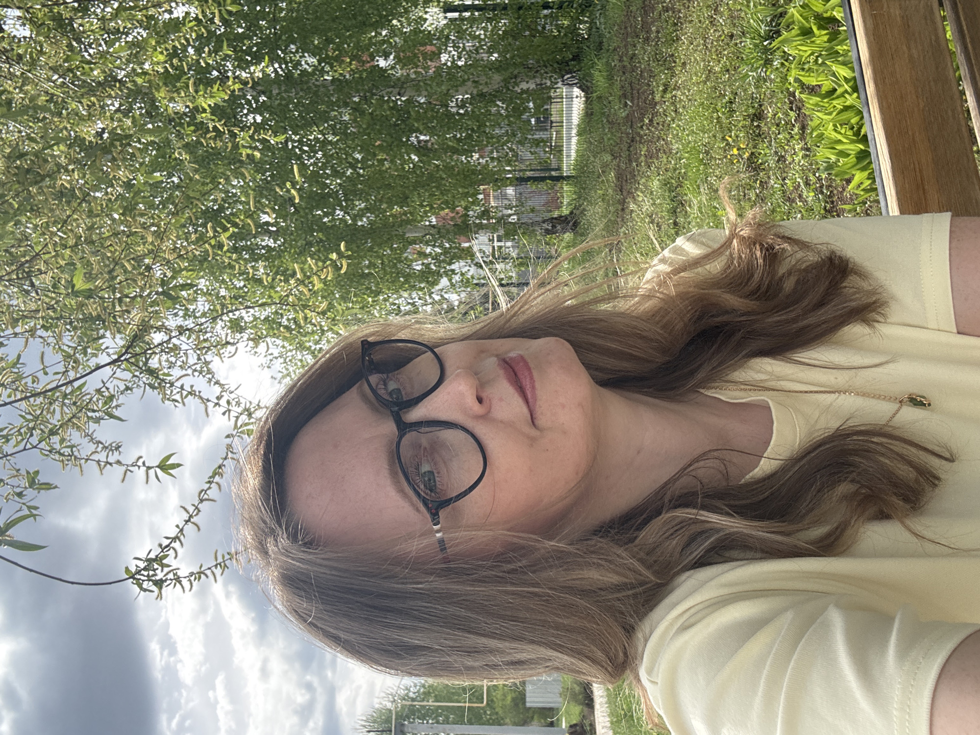 A woman with light brown hair, wearing black-rimmed glasses and a yellow t-shirt, is outdoors. She appears to be sitting on a wooden bench. The background includes green trees and grass, with a white fence visible in the distance. The sky is cloudy. The location is outdoors in Chaikovsky, Russia.