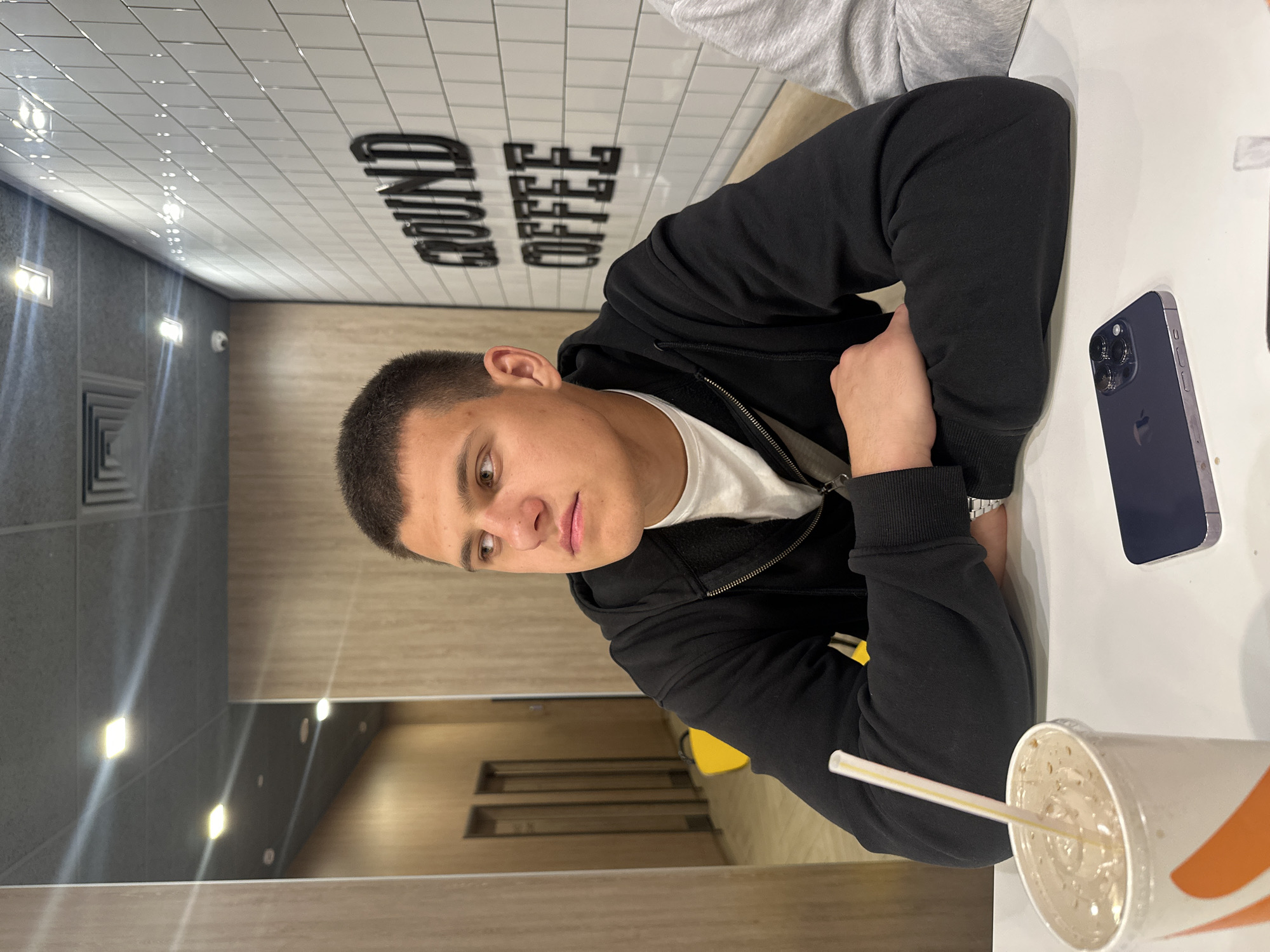 In Birobidzhan, Russia, a young man with short, dark hair is seated at a white table in a cafe, his arms crossed. He is wearing a black hooded sweatshirt over a white t-shirt. To his left, a disposable cup with a straw contains a brown beverage with ice. To his right, a dark-colored smartphone is lying on the table. The cafe's interior features wood-paneled walls and white tiled walls with black lettering that reads "GROUND COFFEE." Overhead, square lights are embedded in a suspended ceiling. In the background, a hallway with doors and yellow chairs is visible.