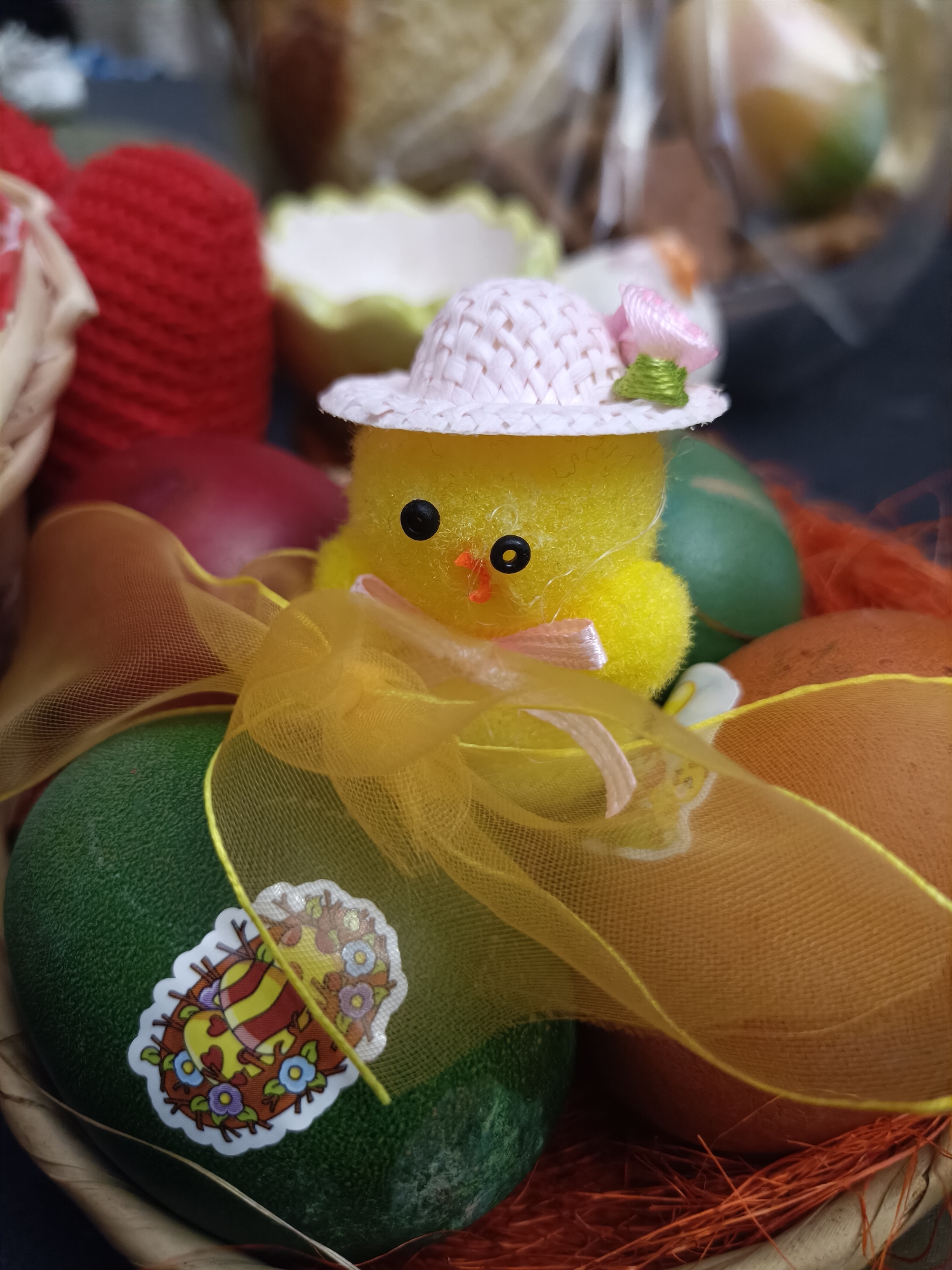 A close-up shot depicts an Easter basket filled with decorated eggs and a small, fuzzy yellow chick wearing a miniature pink hat. The chick is positioned prominently in the center of the frame, surrounded by eggs dyed in green and brown hues. A translucent yellow ribbon is draped around the chick and eggs. One of the green eggs is adorned with a sticker featuring a cartoon image with flowers and stripes. The basket appears to contain brown straw.
The context hints suggest this image may be from Plovdiv, Bulgaria, but there is no visual evidence confirming this. No people or actions are visible within the frame. The focus is on the Easter decorations and the celebratory theme.