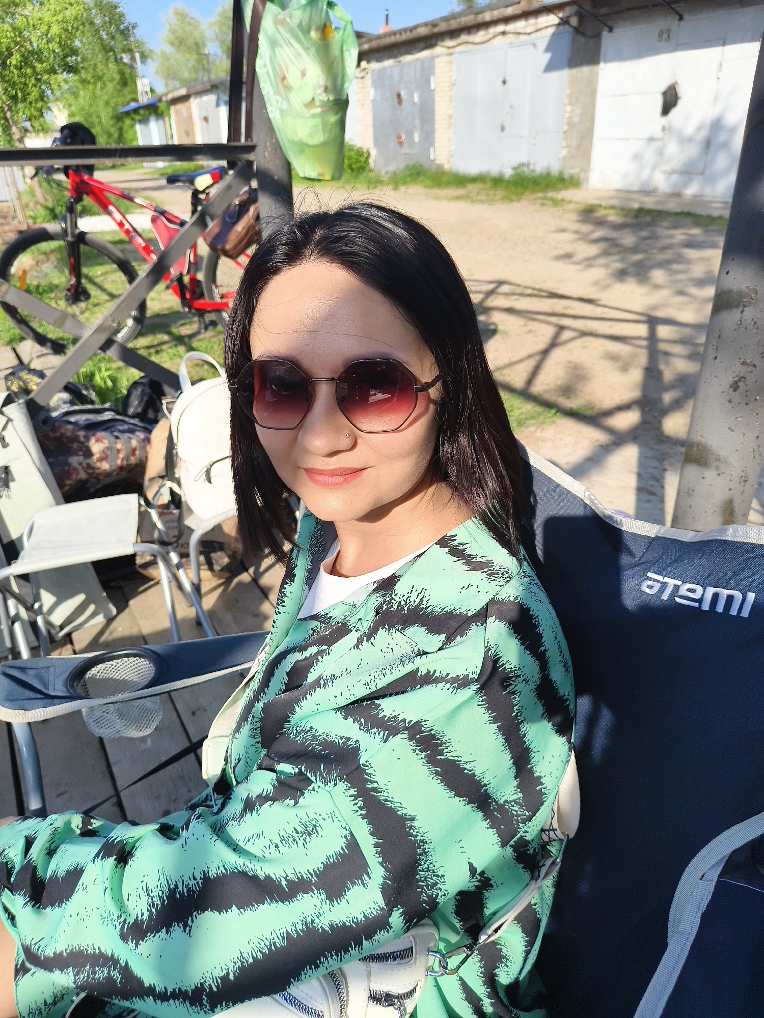 A woman wearing sunglasses and a green and black patterned shirt is sitting in a blue camping chair, outdoors in an urban or suburban area of Unknown, Unknown. Behind her is a red bicycle. In the background, there are several garage structures.