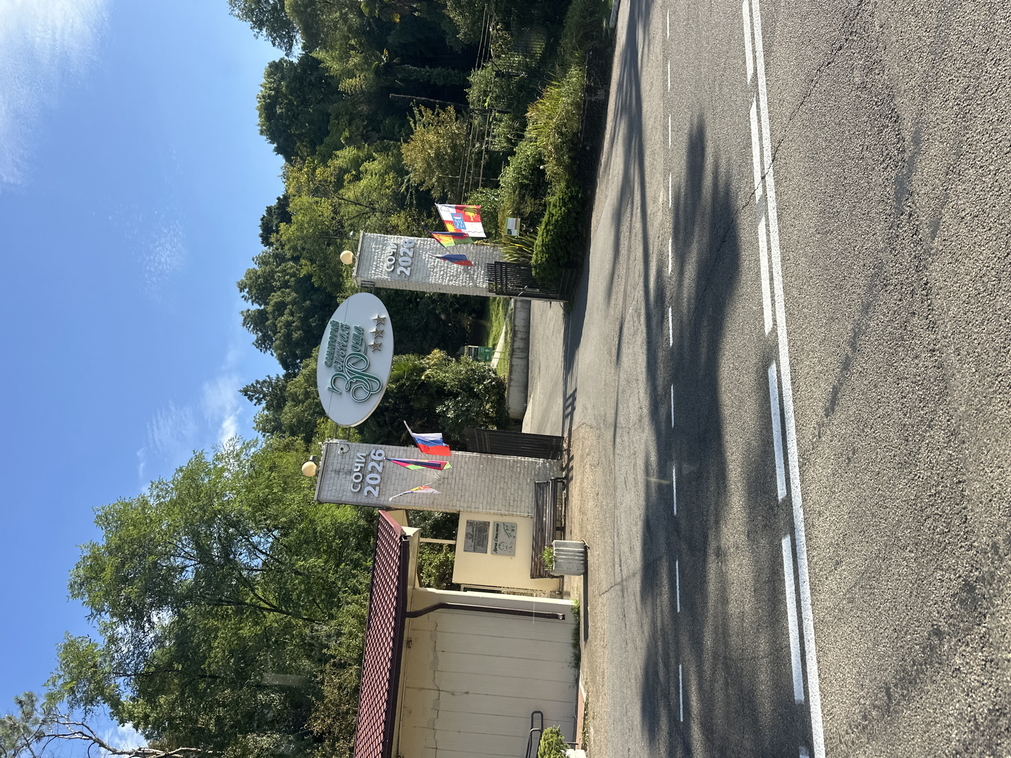 The image shows the entrance to a sanatorium in Sochi, Russia, with a prominent sign featuring the text "СОЧИ 2026" (Sochi 2026) in Cyrillic and a circular logo with the words "САНАТОРИЙ ЗЕЛЕНАЯ РОЩА" (Sanatorium Zelyonaya Roshcha). Several flags are displayed at the entrance. The architecture includes white brick pillars and an attached building with a red tiled roof. A bench is visible near the entrance. The scene is set against a backdrop of lush green trees and a clear blue sky. The foreground shows a paved road with white lane markings. There are no people visible in the image.