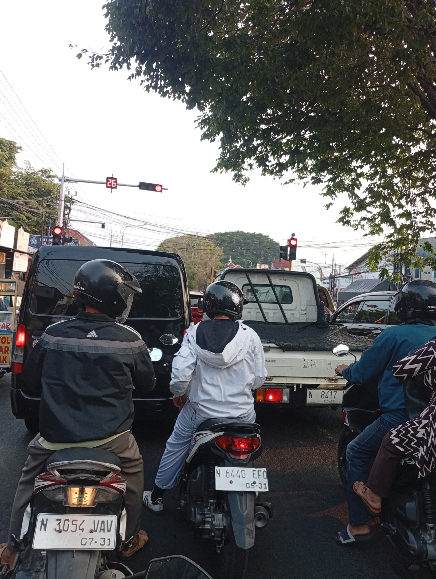 In Singosari, Indonesia, at an intersection on a street lined with trees and power lines, a traffic light displays a red signal with a countdown timer showing "26." Several vehicles are stopped, suggesting a traffic jam. In the foreground, two individuals on motorcycles are visible from behind. The person on the left wears a black jacket with grey stripes and a black helmet. The person on the right wears a white hooded jacket and light blue pants, also with a black helmet. Behind them, a black van is stopped, followed by a white pickup truck. Further back, other vehicles and buildings line the street. The lighting suggests it is either early morning or late afternoon.