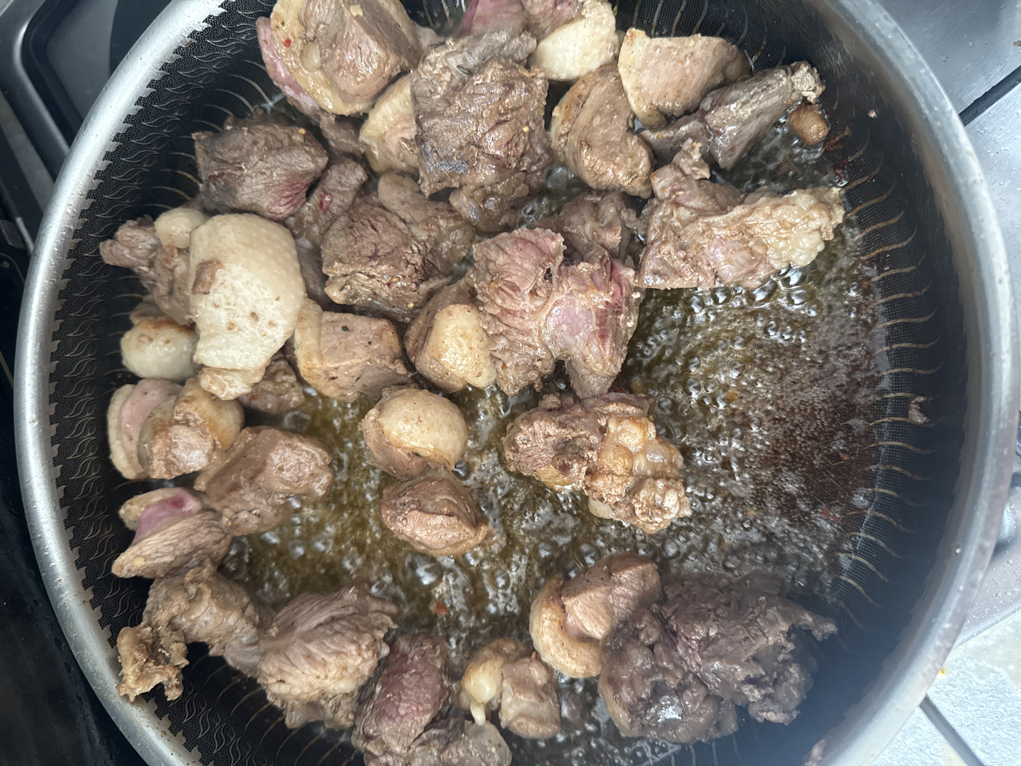 The image shows bite-sized pieces of raw meat, possibly beef or lamb, being pan-fried in hot oil. The meat pieces are irregular in shape and size, with some exhibiting pinkish interiors and browned exteriors from the searing process. The oil in the pan is bubbling vigorously, indicating a high temperature. The pan itself appears to be a non-stick skillet with a textured interior and a metallic rim. The background is blurred, but suggests a kitchen environment, possibly a stovetop. There is no indication of the location being Bishkek City, Kyrgyzstan, or any specific city. The scene is devoid of any people or explicit indicators of human presence other than the act of cooking.