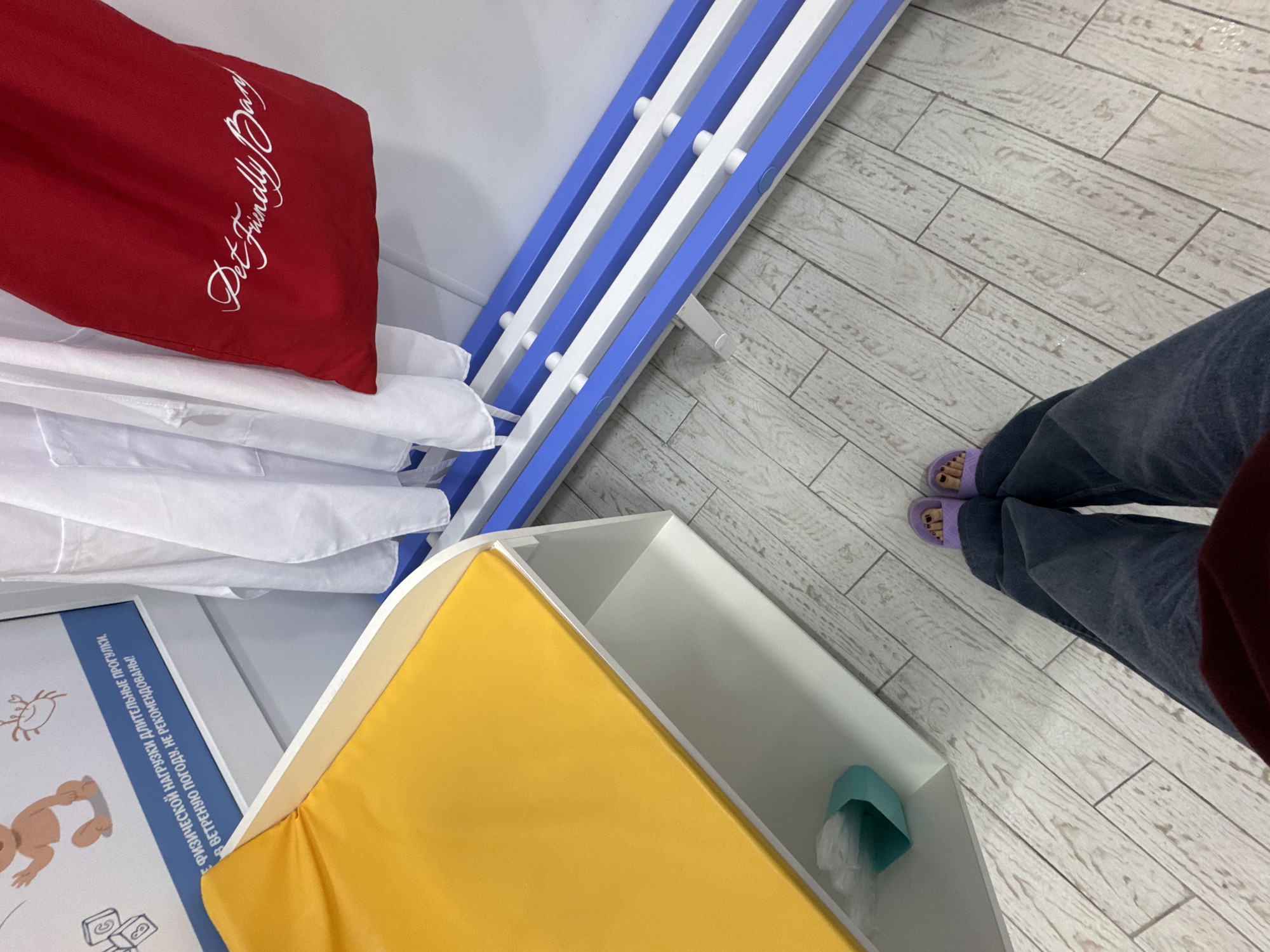 The image captures an indoor scene, likely in a pet-friendly facility or veterinary clinic in Sochi, Russia, viewed from a high-angle perspective. The lower legs and feet of a person wearing dark jeans and purple open-toed sandals are visible on the right, standing on a light-colored, wood-look tiled floor.
In the upper left, a red pillow or cushion with the white embroidered text "Pet Friendly Day" is prominent, resting on a stack of neatly folded white fabrics. Adjacent to this, a blue and white slatted structure extends diagonally across the mid-ground, possibly a bench or drying rack.
The lower left features a partially visible poster with a simple illustration of a dog and other symbols. Accompanying Cyrillic text reads: "ФИЗИЧЕСКОЙ НАГРУЗКИ ДЛИТЕЛЬНЫЕ ПРОГУЛКИ, В ВЕТРЕНУЮ ПОГОДУ, НЕ РЕКОМЕНДОВАНЫ!", which translates to "PHYSICAL EXERTION, LONG WALKS IN WINDY WEATHER, ARE NOT RECOMMENDED!" This advice, along with the "Pet Friendly Day" pillow, indicates a setting focused on animal welfare.
In the bottom center, there's a white-framed structure with a bright yellow padded surface, possibly a changing station or resting area for pets. Next to it, an open white compartment holds a small teal-colored container with plastic inside, possibly a waste bin or dispenser. The overall impression is a clean and organized environment designed for pet care.