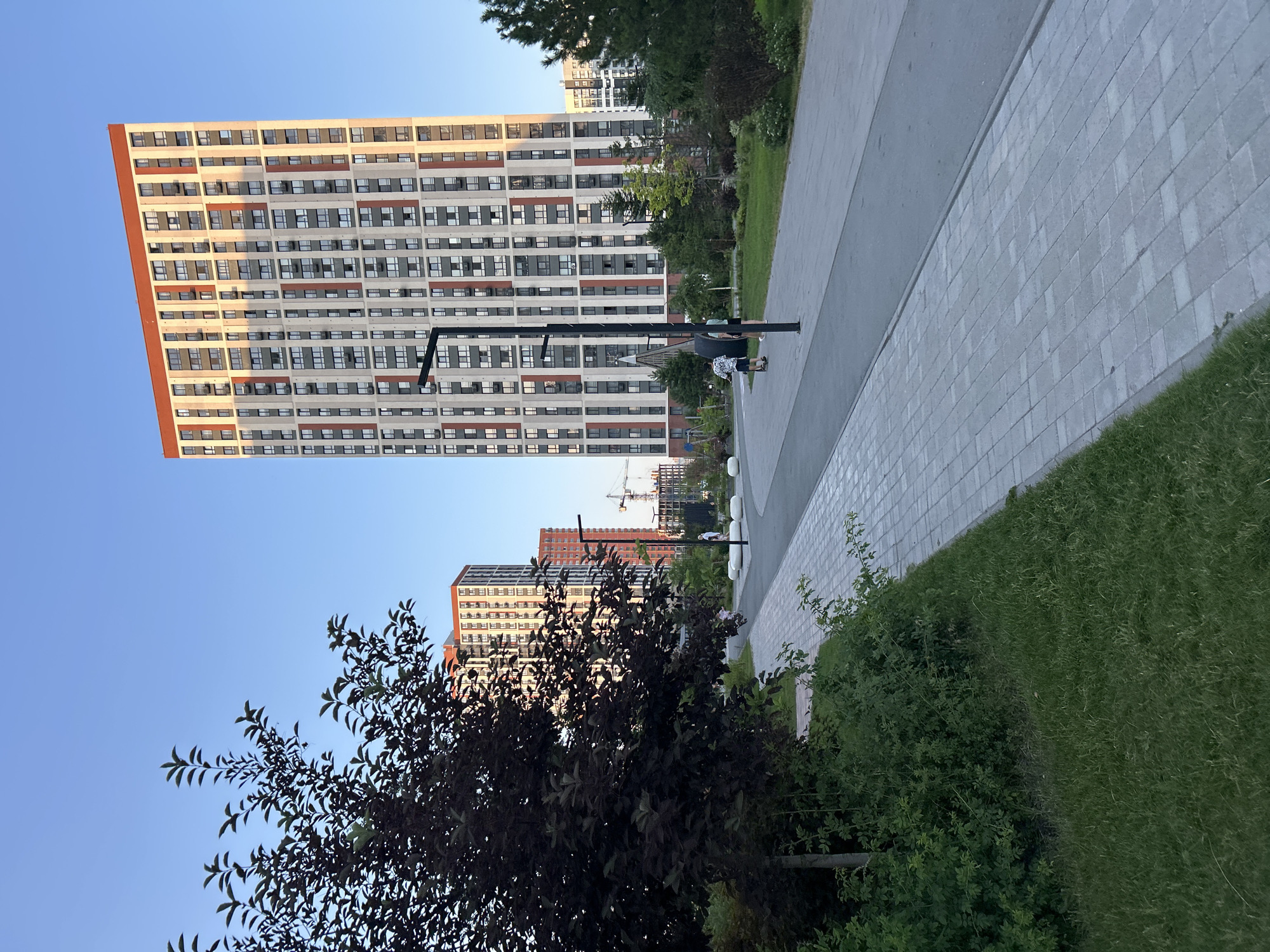 This is a medium shot, taken outdoors, of a modern residential complex with tall buildings. The buildings are predominantly white with orange accents and feature many windows. In the foreground, there is a paved walkway with a grassy area to the right. A dark-colored tree with rich foliage is situated to the left of the walkway, obscuring part of the lower section of the buildings. A streetlamp stands on the walkway near a bench with two people sitting on it. The sky is clear and blue, suggesting a sunny day. The sun appears to be low in the sky, casting long shadows and highlighting the buildings. The overall impression is one of a contemporary urban or suburban environment with well-maintained landscaping. There are no visible text elements in the image.