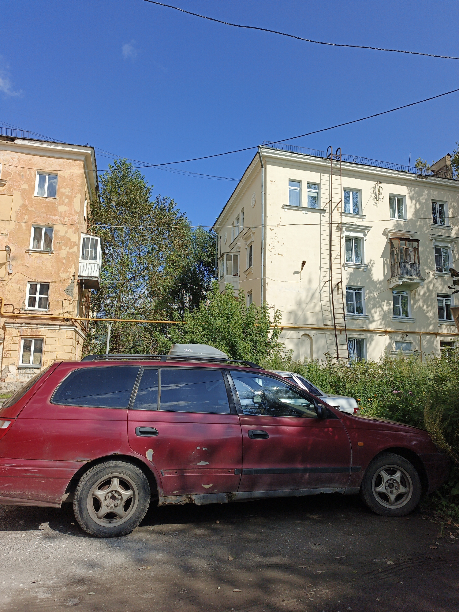 A weathered red station wagon is parked on a gravel lot in front of two apartment buildings. The car has a roof-mounted cargo box and appears to be in a state of disrepair, with rust visible on the lower body panels and door handles. The buildings are typical of Soviet-era architecture, with cream-colored and faded peach facades, multiple windows, and balconies. Green trees and bushes surround the buildings and partially obscure the station wagon. The sky is a clear, bright blue. The scene depicts an urban residential area in Zlatoust, Russia, likely a courtyard or parking area. There are no people or animals visible in the image.