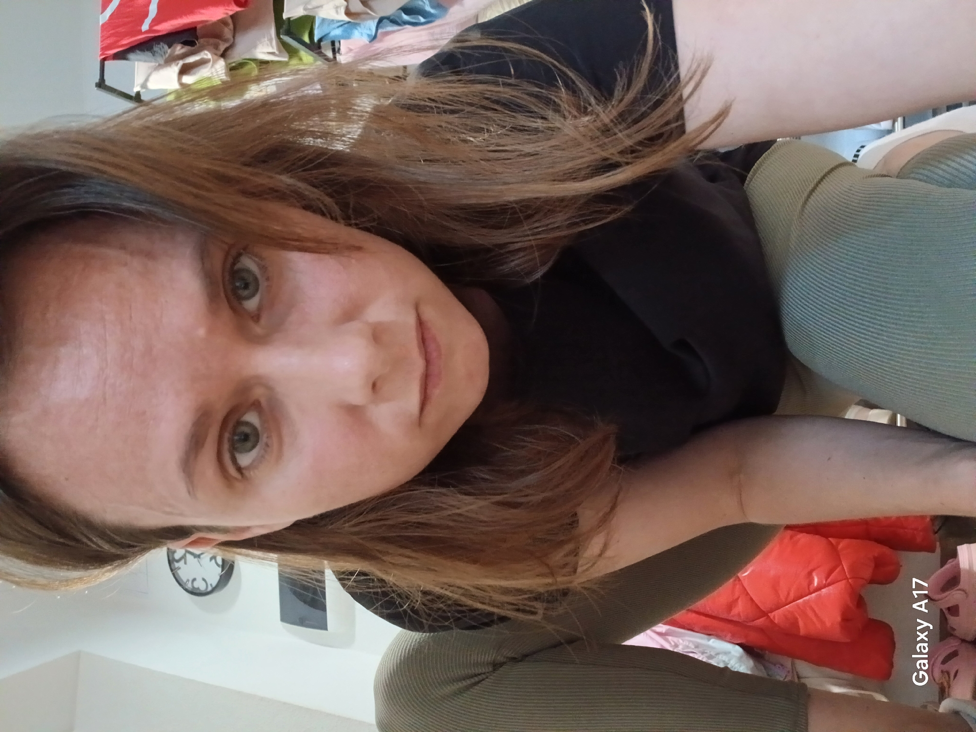 A woman is kneeling, facing the camera. She has light skin, light brown hair, and green eyes. She is wearing a dark shirt and ribbed, olive-green pants. In the background, there is a white wall with a round clock hanging on it, a red puffer jacket, and children's shoes. The phone model "Galaxy A17" is visible in the lower right corner of the image. The location is likely indoors, but there is no specific indication of Yekaterinburg, Russia.