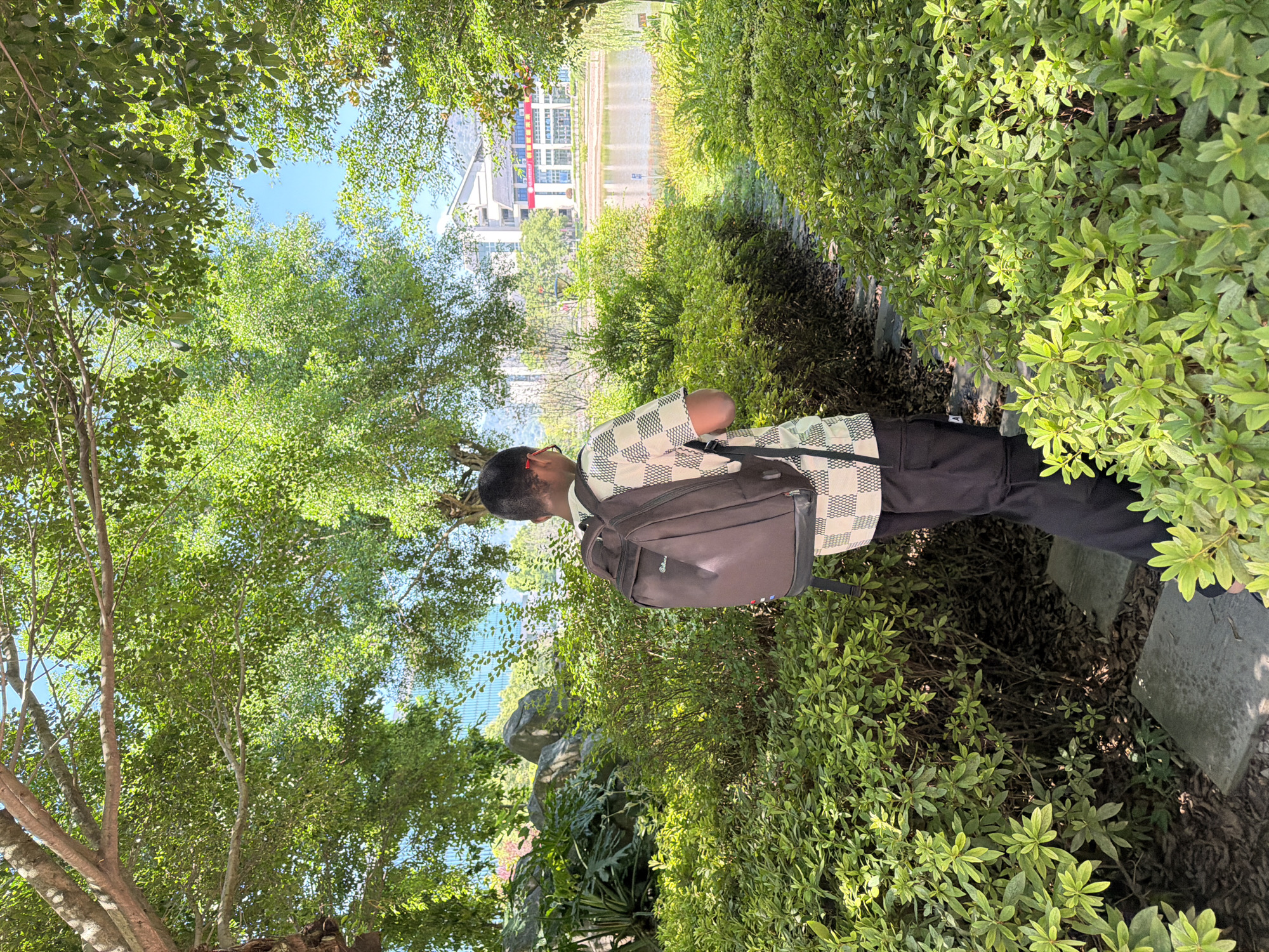 A young person wearing glasses, a checkered shirt, and black pants walks along a stone path through a lush green garden in Jiangjin, China. The person carries a dark brown backpack. Dense foliage surrounds the path, with large trees overhead dappling the scene with sunlight. In the background, a body of water reflects the sky and a modern building with a red sign is visible. The overall atmosphere is serene and natural.