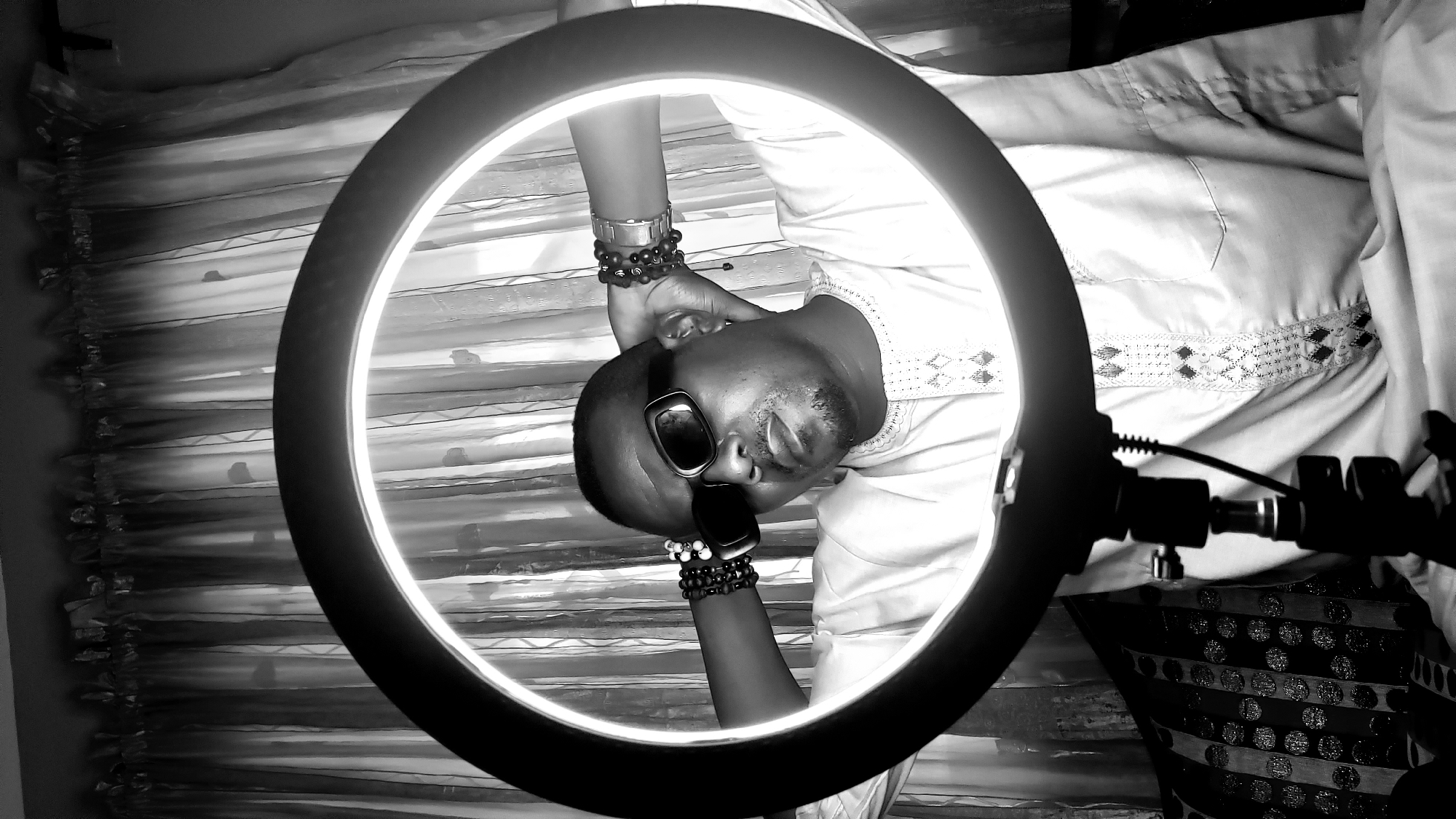 A person wearing sunglasses and a beaded bracelet stands in front of a window with sheer curtains. A ring light illuminates the person's face and upper body. The location is Narayi, Nigeria. The image is in black and white.