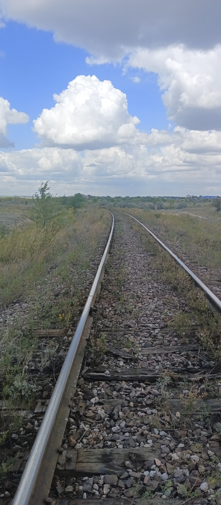 The image depicts a set of railroad tracks curving to the right, disappearing into the distance. The tracks are laid on a bed of gravel and weathered wooden sleepers. The surrounding environment is a dry, grassy landscape with sparse vegetation and some scattered bushes. In the background, a distant town or settlement is visible, characterized by low-rise buildings. The sky is partly cloudy, with large, fluffy white cumulus clouds set against a bright blue sky. The overall impression is one of a rural or semi-rural area, possibly on the outskirts of a city. The specific location in Karaganda, Kazakhstan, is not discernible from the visual information alone.