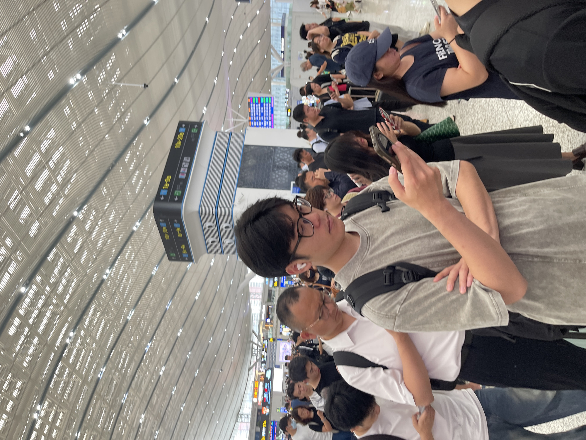 This is a photograph taken in a brightly lit, modern indoor space that appears to be an airport or train station in China. The ceiling is a prominent feature, with a complex, arched design of white panels and integrated lighting, creating a grid-like pattern that stretches into the distance.
The scene is populated by a crowd of people, many of whom are standing or sitting, looking at their phones or waiting. The people are diverse in age and attire, suggesting a busy travel hub. In the foreground, a young man with glasses is looking towards the camera, holding a smartphone in his hand. To his left, an older man in glasses is also looking down. Further back, more individuals and groups are visible, some with luggage.
Several signs are visible in the background, some displaying what appear to be departure or arrival information in a combination of Chinese characters and numbers, such as "10-20e" and "1E-9e". The overall atmosphere seems to be one of waiting and transit, with no strong indication of emotion, but a general sense of busyness. The lighting suggests it is daytime, and the indoor setting implies the weather outside is not a factor. The architectural style and signage point to a contemporary transportation facility in China, likely within Longhua District.