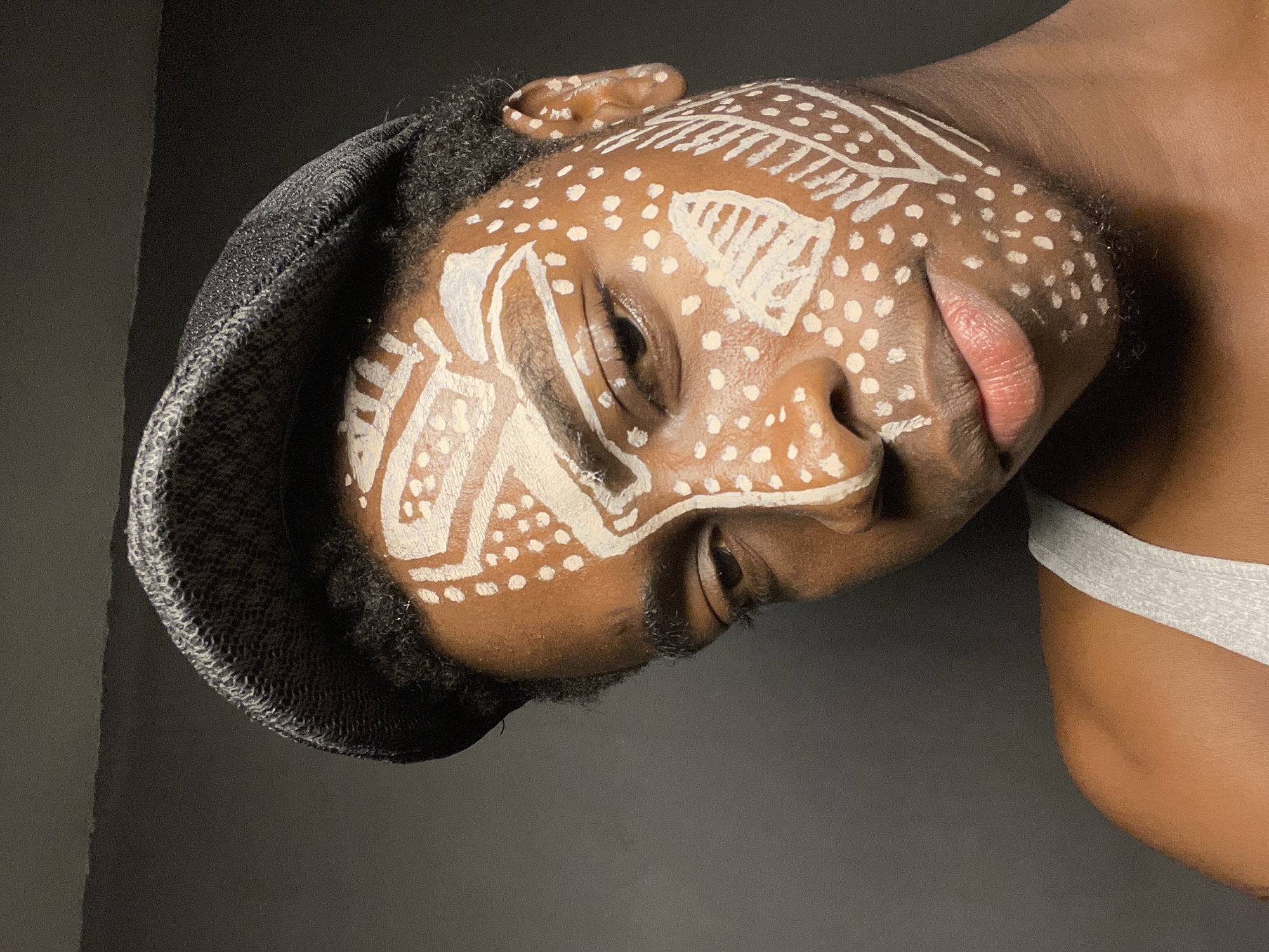 A close-up, medium shot depicts a Black person with white paint on their face, in a style reminiscent of tribal markings, with a pattern of dots and lines. The person wears a gray cap and a gray tank top is visible on their shoulder. The background is dark gray. The location in Nkpor, Nigeria cannot be determined from the image.