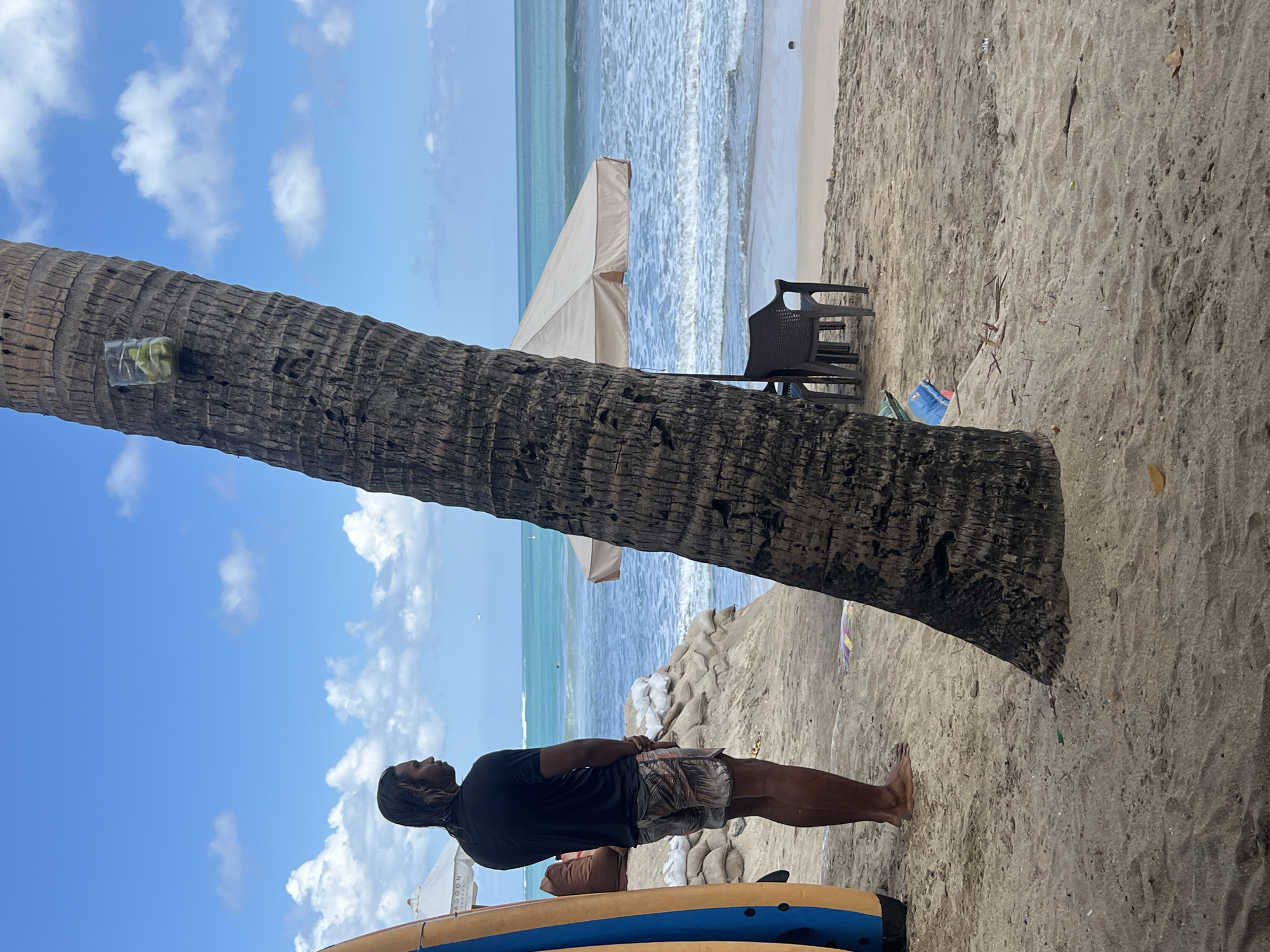 In Kuta, Indonesia, a person stands on a sandy beach, looking out at the ocean. They are wearing a black t-shirt and patterned shorts. To their left, a surfboard leans against a palm tree. Sandbags are piled up behind them, suggesting a potential barrier against the sea. In the background, the turquoise ocean meets a pale blue sky with scattered white clouds. A beige umbrella shades two dark plastic chairs on the sand. The foreground is dominated by the textured trunk of a palm tree, with a clear plastic cup containing slices of lime affixed to it. The sand is strewn with small debris and dried leaves.