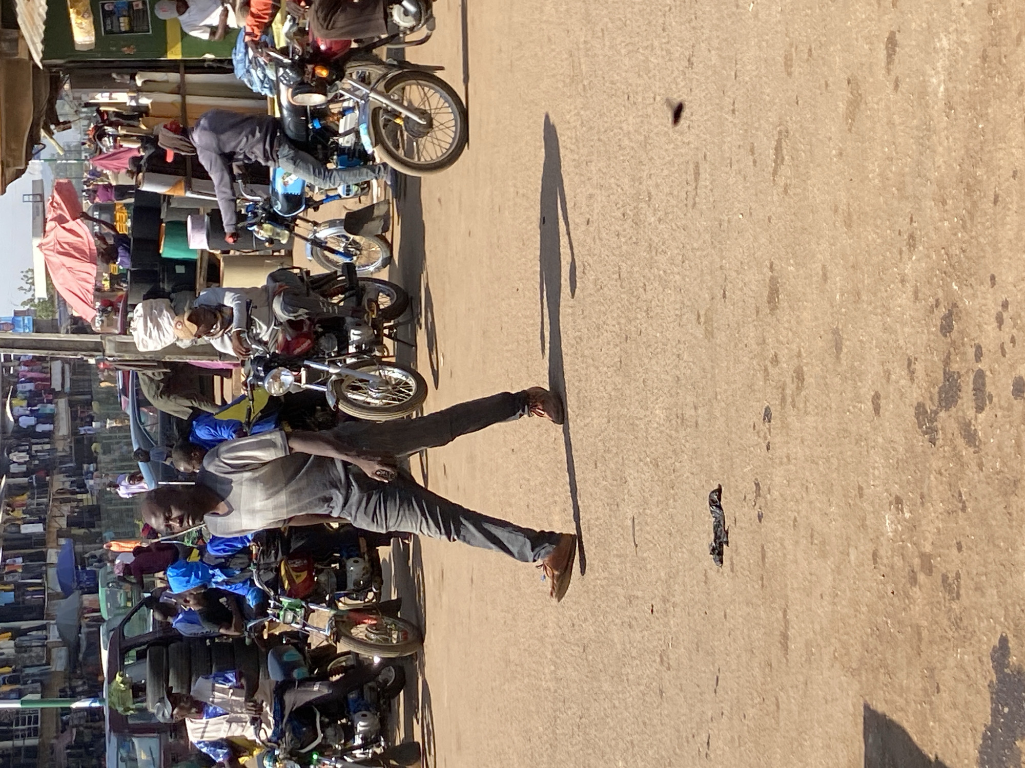 The image shows a street scene in Bukuru, Nigeria, during daylight. A man in a grey striped shirt and dark pants is walking across a paved road, his shadow cast beside him. To his left are several parked motorcycles, some with riders or passengers. In the background, a densely populated area with shops or stalls is visible, with various items displayed under awnings and umbrellas. A vehicle is also partially visible among the motorcycles. The road surface is slightly uneven and worn.