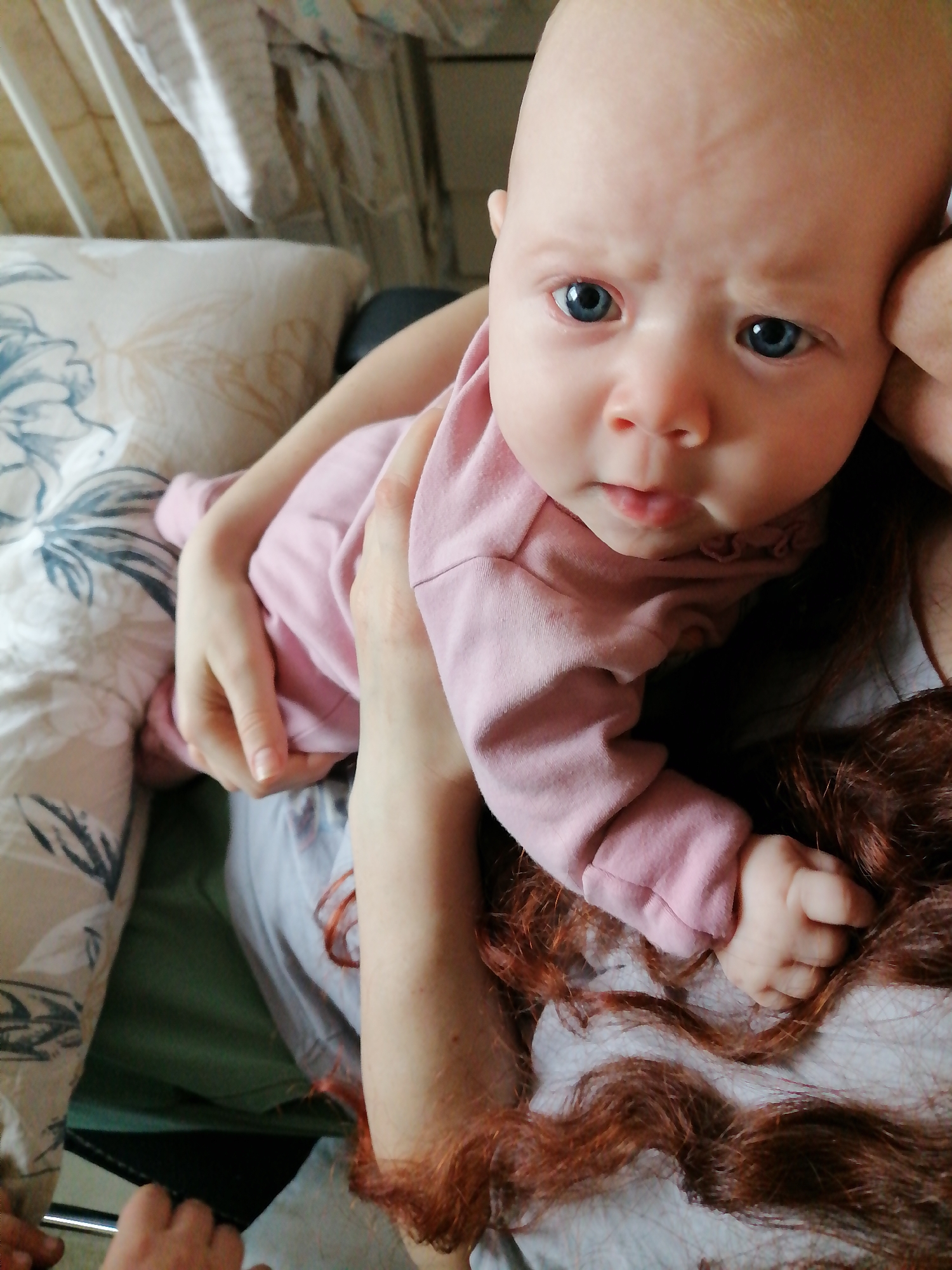 A baby is being held in someone's arms. The baby has light skin, blue eyes, and is wearing a pink outfit. The adult holding the baby has red hair. There is a pillow with a floral pattern to the left of the baby. The setting appears to be indoors, possibly in a home.