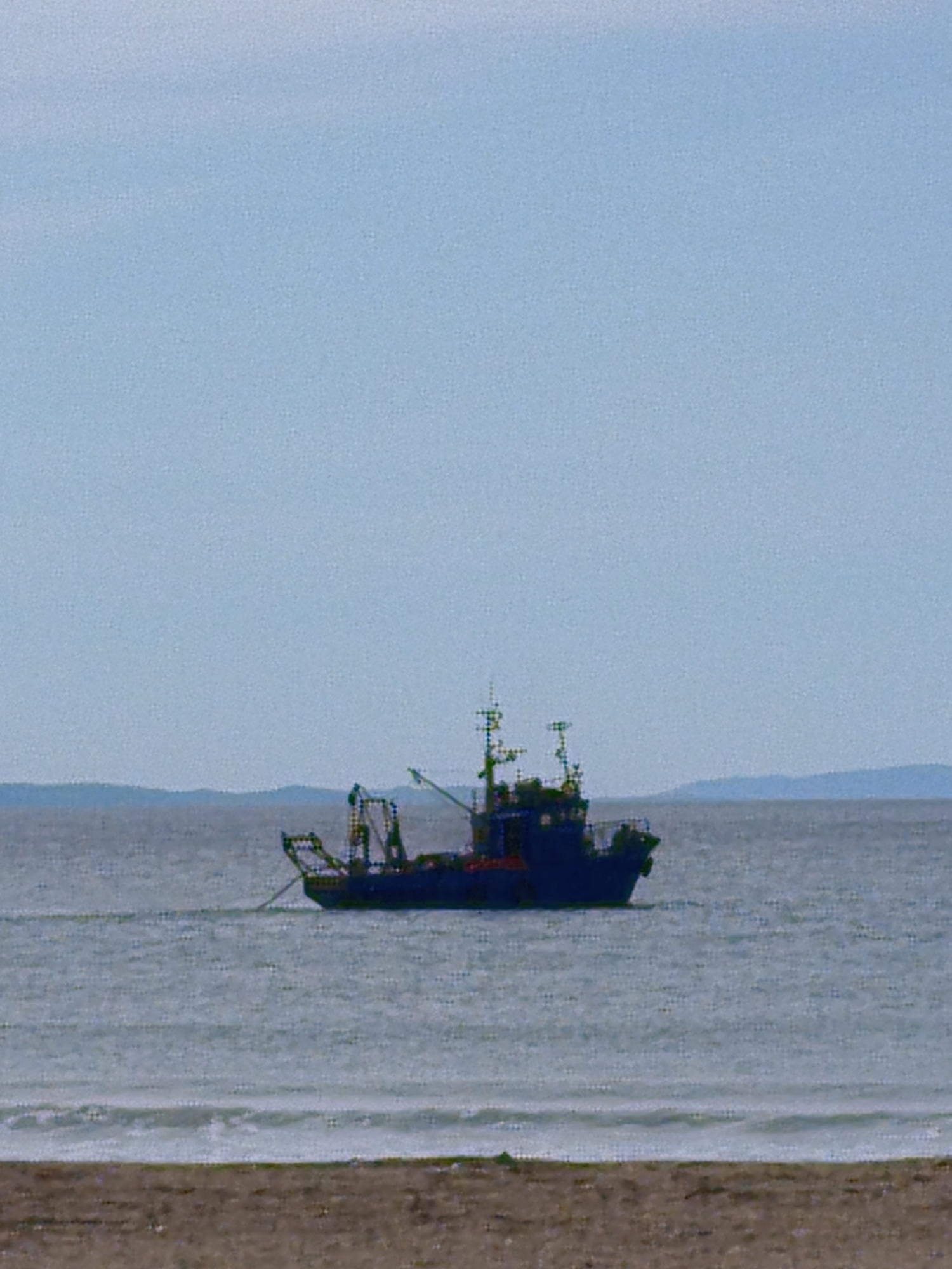A fishing trawler is sailing offshore near a beach. The sky is pale blue with slight haze. The sea is pale gray with small waves. The boat is dark blue and sits in the middle of the image, facing to the right. It has multiple masts and booms. The beach is in the foreground, colored brown and gray. There are low, rolling hills in the background, which are pale blue-gray. The location is unknown.