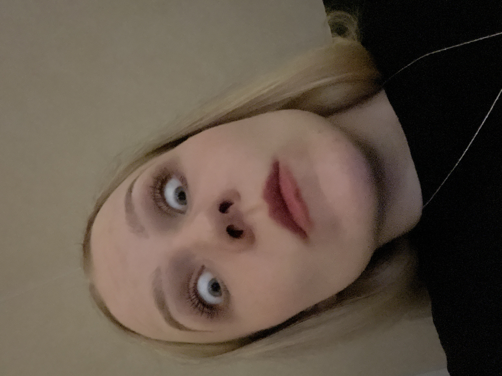 This close-up portrait, oriented sideways, features a young woman with fair skin and blonde hair. Her face is the central focus, looking directly at the viewer with wide, light-colored eyes. Her eyebrows are prominent, and her eyelashes appear long and dark. Her lips are slightly parted, revealing a natural or applied reddish-pink color.
She is wearing a dark, possibly black, garment, a portion of which is visible on the right side of the frame. The background consists primarily of a plain, solid light brown or beige surface, likely an interior wall or ceiling. Some strands of her blonde hair are visible around her face.
The lighting is soft and even, suggesting an indoor setting without strong directional light. Her expression is neutral and direct. There are no discernible objects beyond the woman and her clothing, nor are there any visible texts or specific location cues within the image itself. The time of day and weather conditions cannot be determined.