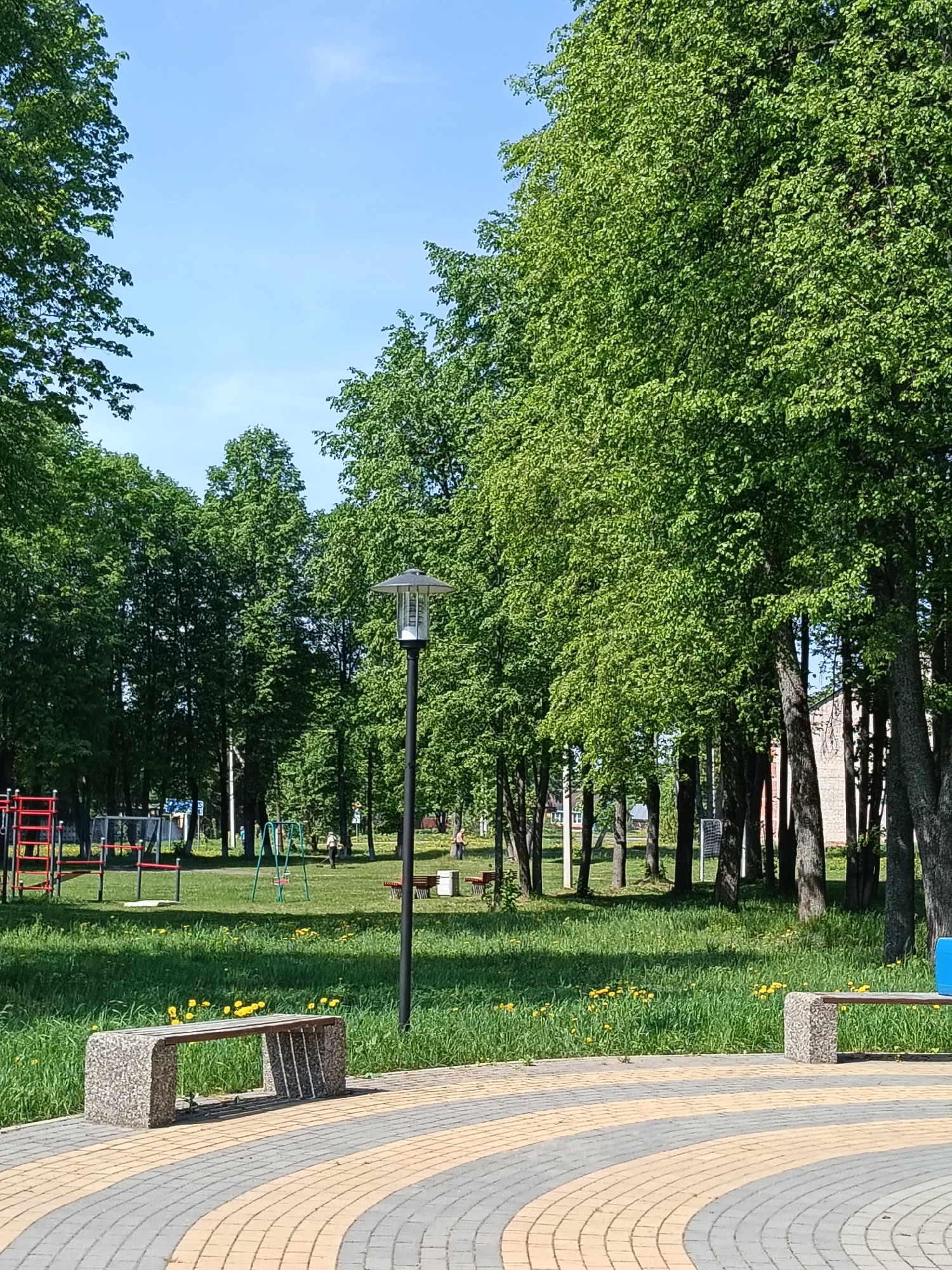 The image depicts a park scene in Красный Маяк, Russia, characterized by a paved area in the foreground featuring a curved pattern of alternating grey and yellow paving stones. Two identical benches, constructed of light grey speckled stone with wooden slats, are positioned on either side of the paved area, facing the lawn.
Beyond the paved area, a grassy lawn extends, dotted with yellow dandelions. In the background are numerous trees with full, green foliage, creating a shaded area. A black lamppost stands in the middle ground, its design modern and simple. A playground is visible in the distance, complete with red and silver metal structures, swings, and what appears to be a basketball court.
Several people are present in the background, mostly individuals casually walking or standing near the playground and benches. They appear to be interacting with the environment, suggesting a relaxed and leisurely atmosphere. The weather is sunny, as indicated by the clear blue sky visible between the trees and the distinct shadows cast in the foreground.