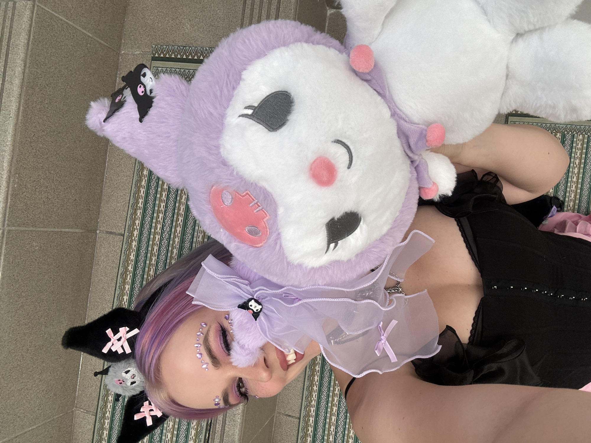The image is a selfie of a young woman lying on a rug with a striped pattern. She is wearing a black corset with lace trim and a pale pink tutu underneath. She has purple hair and is wearing cat ears with pink bows and a small Kuromi plush attached to one of the ears. She has glittery eye makeup and gem accents on her face. She is also wearing a large Kuromi plush hat with a tulle collar. The background shows a tiled floor and a portion of a patterned rug.
The location in the city of City of Zagreb, Croatia is not evident in the image.