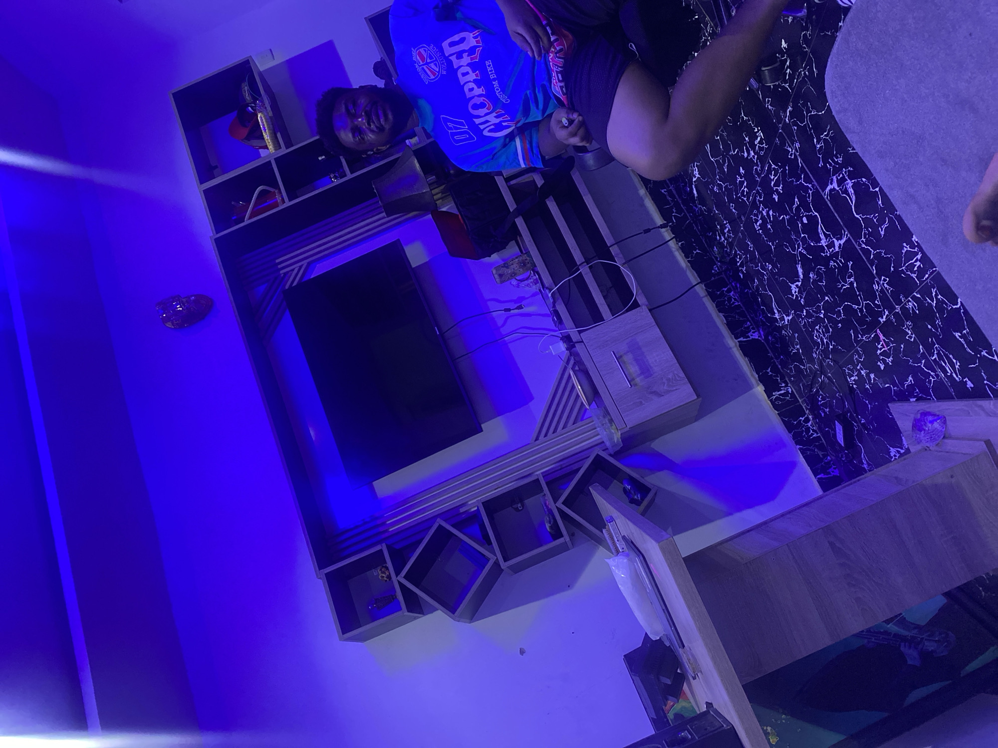 A man is in a room in Abuja, Nigeria. He is wearing a blue shirt and black shorts, and he is sitting on a piece of furniture. Behind him is a wall-mounted television. The room is lit with blue lights. The floor appears to be tiled. In the background, there is a wooden cabinet.
