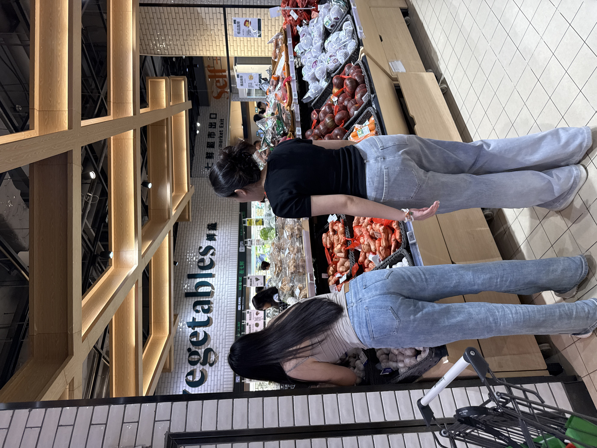 In a brightly lit supermarket in Haizhu District, China, two women are seen shopping for produce. The woman on the left, with long black hair, wears a light-colored tank top and light-wash flared jeans. She is bent over, looking into a basket of garlic. To her left, a shopping cart with a green bag in it is partially visible.
The woman on the right, with her dark hair pulled up, wears a black t-shirt and similar light-wash flared jeans. She stands facing away from the camera, observing the vegetable displays. In the background, a sign reads "Vegetables" in English and Chinese characters. The shelves are stocked with various fresh produce, including red onions, sweet potatoes, and a selection of fruits wrapped in netting. The floor is tiled in a grid pattern, and wooden beams with integrated lighting create an overhead structure. The overall environment suggests a modern, well-organized grocery store.