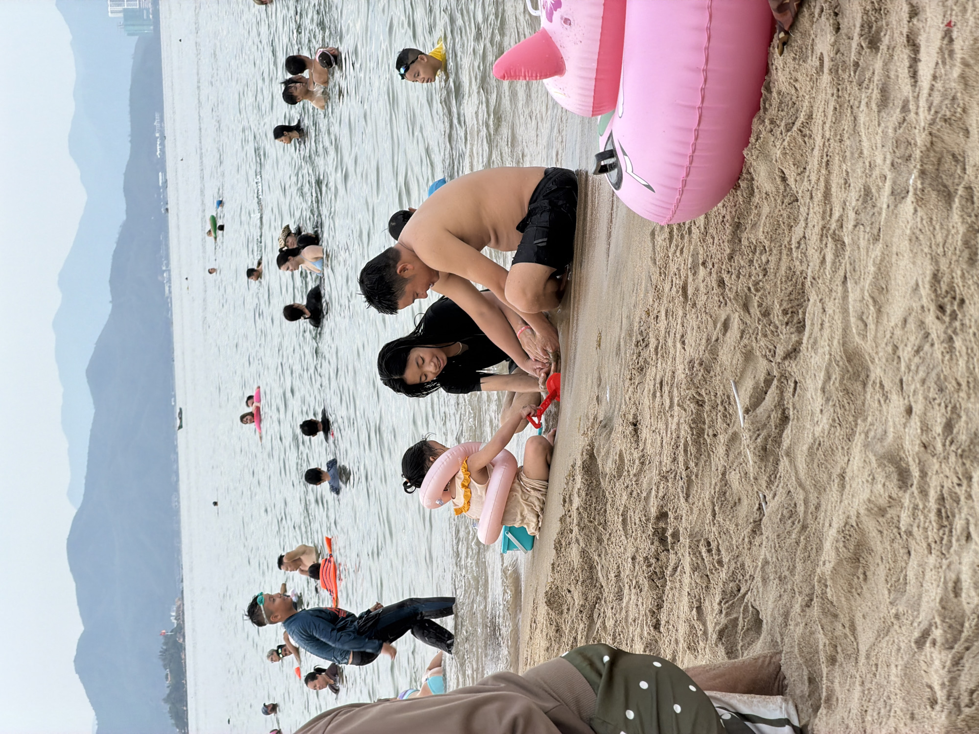 A group of people are at the beach in Nha Trang Ward, Vietnam. In the foreground, a family is playing in the sand and shallow water. A man in black swim trunks sits on the sand, facing a young girl wearing a light-colored dress and a pink inflatable ring around her waist. The girl is playing with a red shovel and bucket. To the right of the girl, a woman in a black t-shirt sits on the sand, looking at the girl. Further back, a boy in a blue shirt and swimming goggles stands in the water. In the middle ground, many other people are in the water, some with inflatable rings, enjoying the sea. The background shows a hazy view of mountains and a distant city skyline. The water is calm and the sky is overcast. In the far right of the foreground, a portion of a large pink inflatable ring is visible on the sand.