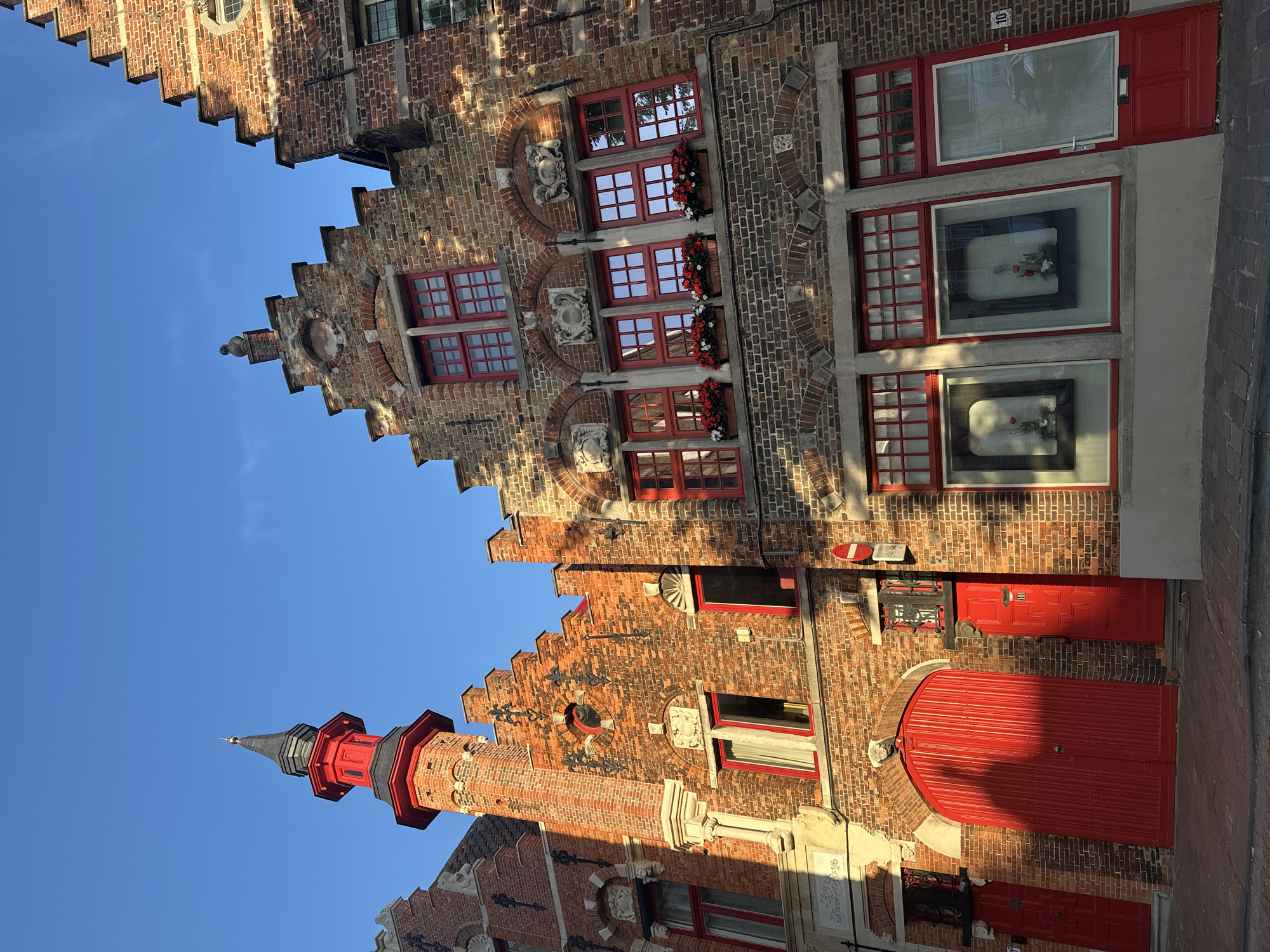The image captures a street scene in Bruges, Belgium, featuring traditional brick buildings with red-trimmed windows and doors under a clear blue sky. The architecture is characterized by stepped gables, ornate decorative elements above windows, and a tall, narrow tower with a red spire on the left building. Large red doors are visible on the ground floor, with one appearing to be a carriage entrance and others serving as building entrances. Flower boxes with red and white blooms adorn some of the windowsills. The sunlight casts long shadows across the brickwork and the paved street. The overall impression is of a well-preserved historical district.