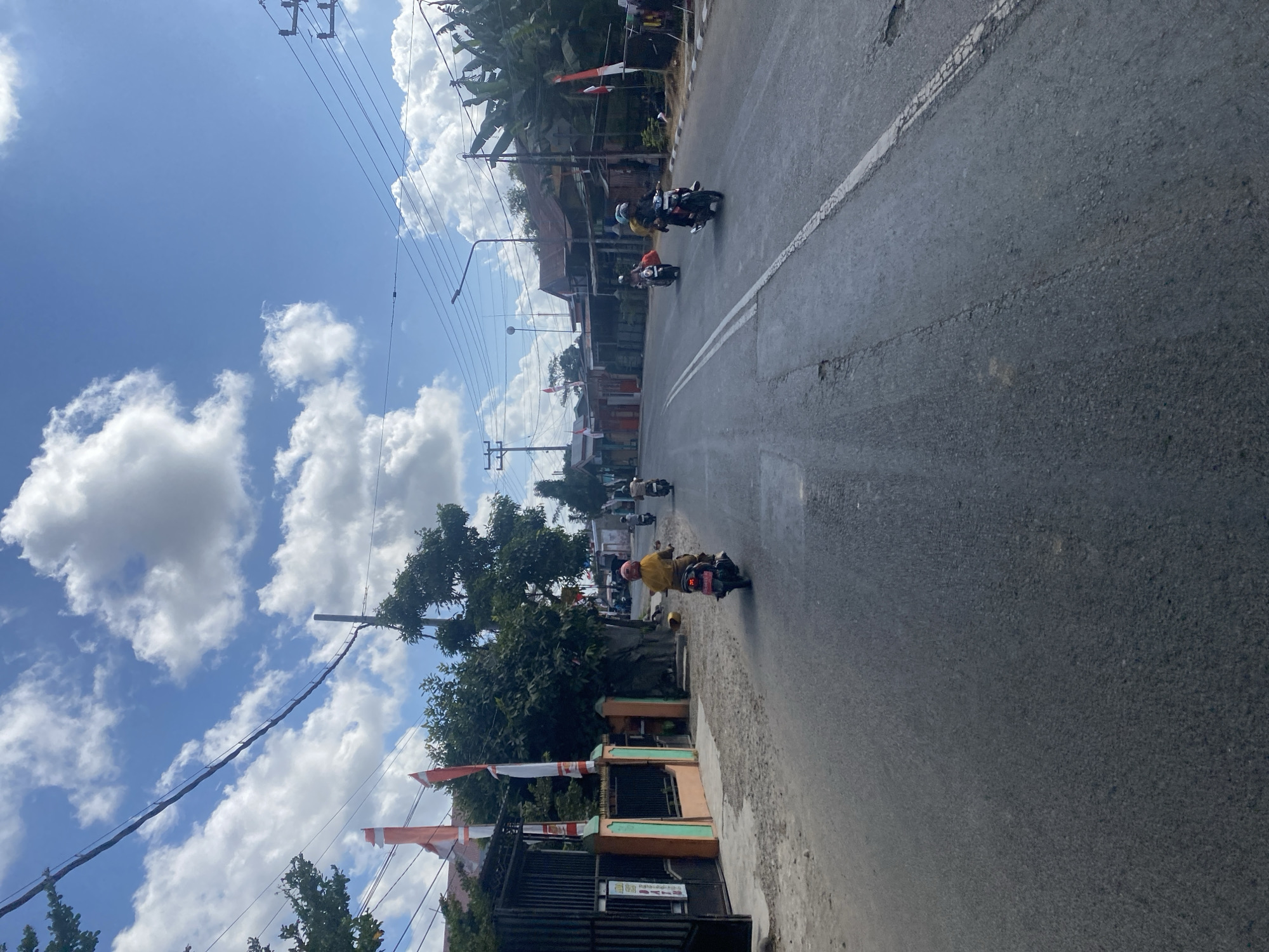 The image depicts a street scene in Bentengnge, Indonesia, under a bright blue sky with scattered white clouds. Several individuals are riding motorcycles on a paved road. One person in the foreground is wearing a yellow shirt and a pink helmet, riding a motorcycle. Further down the road, other motorcyclists are visible, some with passengers. On the left side of the road, there's a gate with an orange and green fence and several Indonesian flags displayed. Trees and buildings line the sides of the street. Utility poles with wires are also present. The road appears to have some wear and tear, with visible cracks and patches. The overall environment suggests a moderately populated area within the city.
