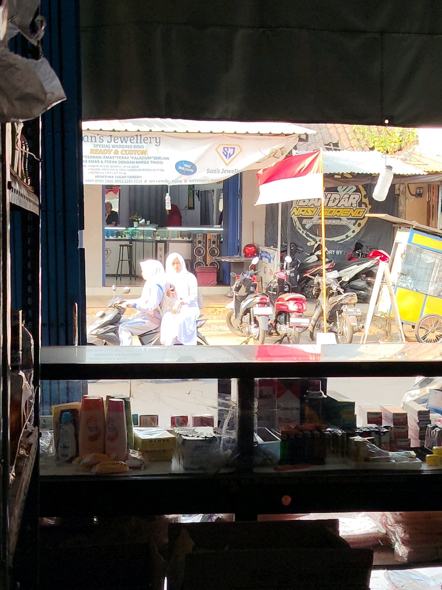 The image depicts a street scene in Dampit, Indonesia, viewed through a darkened interior space, likely a shop. In the foreground, a shelf displays various consumer goods, including shampoo bottles, packaged snacks, and boxes of cigarettes. The mid-ground reveals an outdoor commercial area with several motorcycles parked. Two women wearing hijabs are seated on one of the motorcycles. Behind them, a storefront displays signage for "San's Jewellery" and "Bandar Nasi Goreng," indicating businesses selling jewelry and food. A red and white Indonesian flag is prominently displayed. The background shows buildings and rooftops under daylight conditions. The overall impression is of a bustling local market or commercial street.