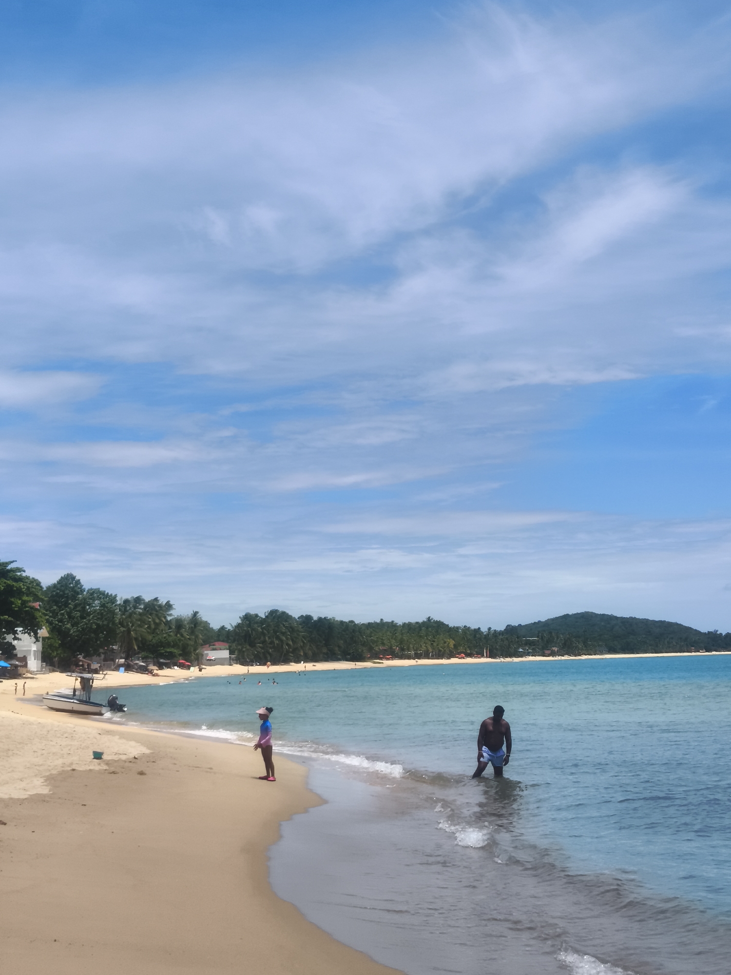 The image depicts a tropical beach scene in the Ko Samui District of Thailand. The foreground is dominated by a wide expanse of golden sand leading to the ocean. To the left, a small white boat with an outboard motor is beached on the sand. Further left, a cluster of buildings and lush green trees form the backdrop. In the mid-ground, a young person wearing a pink and blue swimsuit stands near the water's edge, facing away from the camera. Further out in the shallow, turquoise water, a dark-skinned man wearing blue swim trunks stands with his legs submerged. The ocean extends to the horizon, where it meets a line of palm trees and a distant, tree-covered hill under a bright blue sky dotted with wispy white clouds. The overall scene conveys a relaxed, sunny day at the beach.