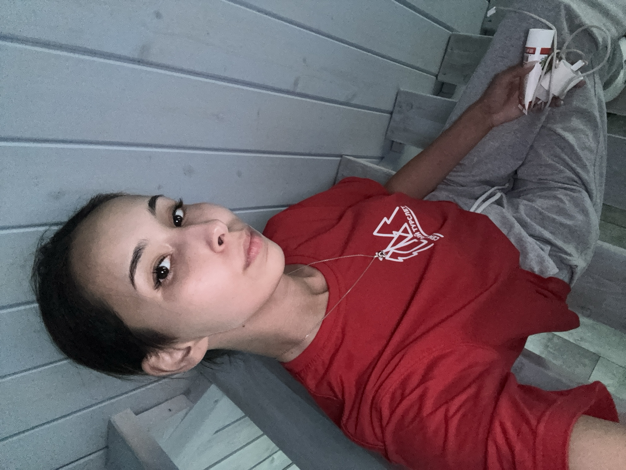 A young woman is seated on a wooden staircase, possibly outdoors, given the natural light and the unfinished wooden structure of the stairs and walls. She is wearing a red t-shirt with a white graphic and a silver necklace, and grey sweatpants. She is holding a small white cylindrical object with red markings in her right hand. Her hair is dark and pulled back, and she is looking directly at the camera. The background consists of horizontally paneled light blue wooden walls and the grey wooden steps of the staircase. The image appears to have been taken from a high angle. The location is identified as Doma 1096 Km, Russia.