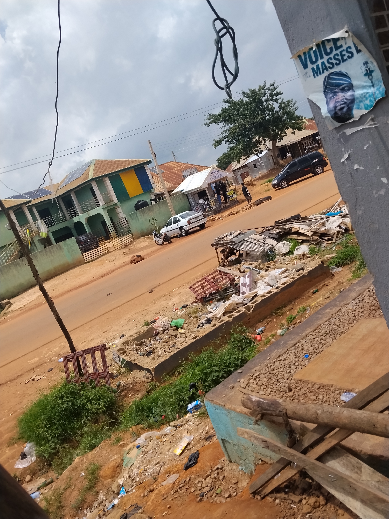 A street scene in Durumi, Nigeria, captured from a slightly elevated perspective looking down and across the road. A dirt road runs through the center of the frame, with a green building with yellow and blue accents on the left, featuring solar panels on its roof and a balcony. A white car and a motorcycle are parked on the side of the road near the building. Across the street, a black car is visible, and beyond that, several tin-roofed structures and trees form the background. A poster with a picture of a man and text "VOICE the MASSES" is attached to a concrete pole on the right side of the image. The foreground on the right shows a rough concrete structure and wooden planks, with debris and scattered stones on the ground. The left side of the foreground is dominated by green vegetation, with more debris and scattered items, including plastic bags and bottles, on the ground. The sky above is cloudy.