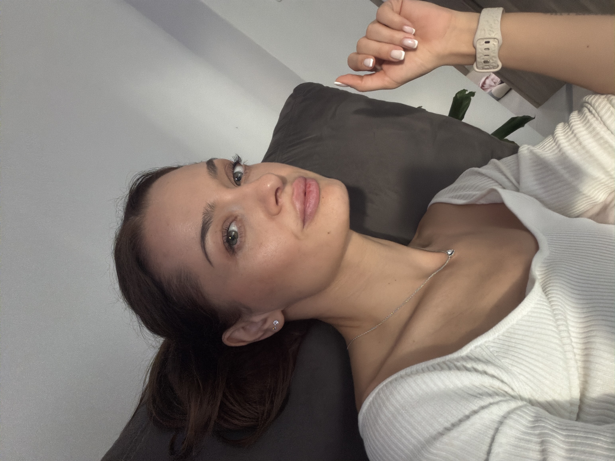 A woman lies on a brown pillow against a light gray wall, wearing a white ribbed top and a silver necklace. Her dark hair is partially pulled back. She is looking up at the camera with a slight smile. Her right hand is raised near her head, displaying a white wristwatch and French-manicured nails. A plant is visible behind the pillow and hand. The image does not include any information that the photo was taken in Varna, Bulgaria.