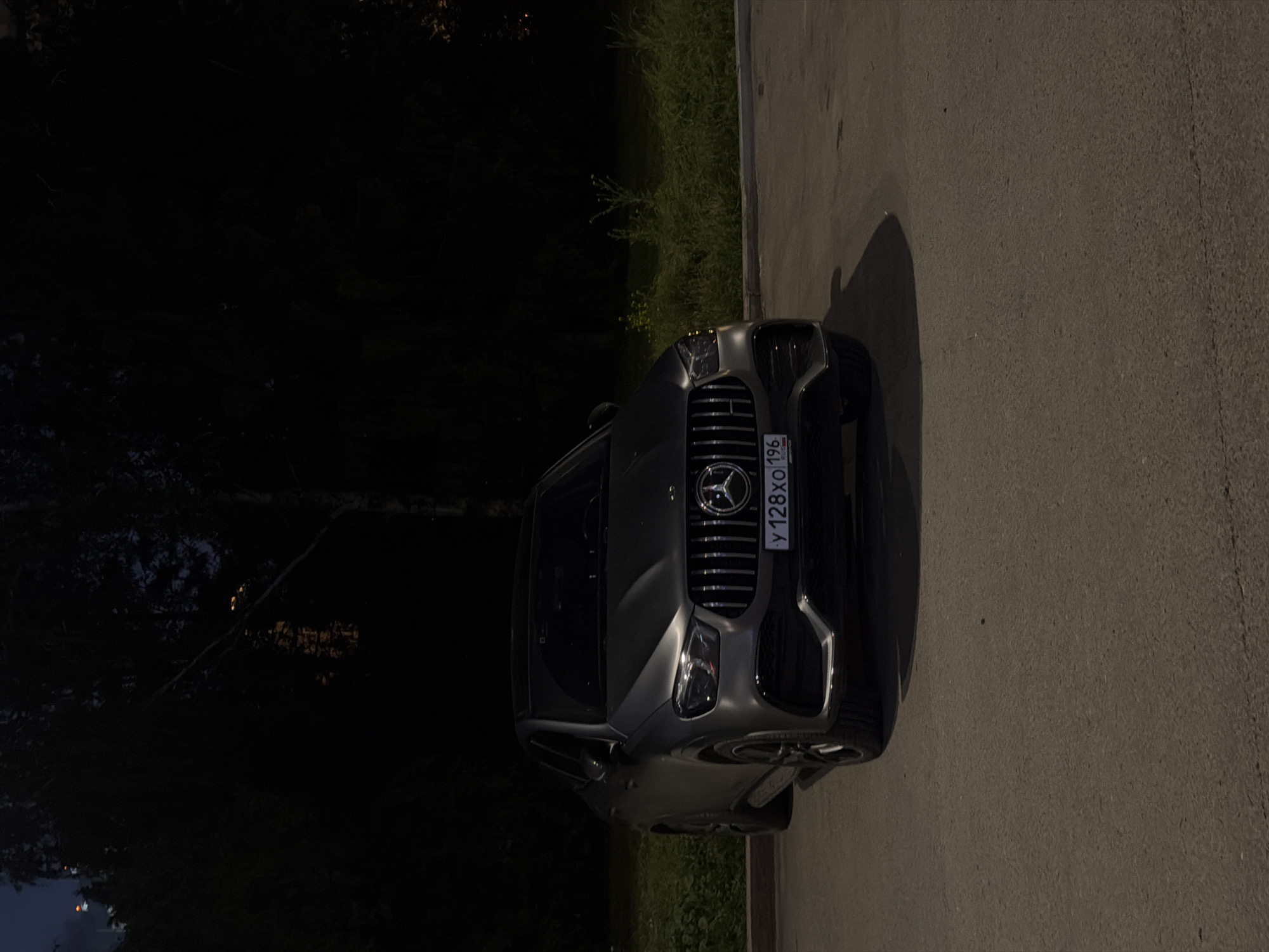 At nighttime, a dark gray Mercedes-Benz GLE Coupe is parked on an asphalt surface. The vehicle is angled slightly towards the viewer's left, with its front grille, headlights, and license plate clearly visible. The license plate displays the alphanumeric characters "y 128xo 196" in white lettering on a dark background, with "RUS" in smaller text below. The car is positioned in front of a line of dense, dark green foliage and trees, which obscure the background. The overall lighting suggests that the scene is illuminated by artificial light sources, possibly streetlights or lights from nearby buildings, creating a contrast between the illuminated car and the dark environment. The asphalt surface appears to be a parking lot or a quiet street.