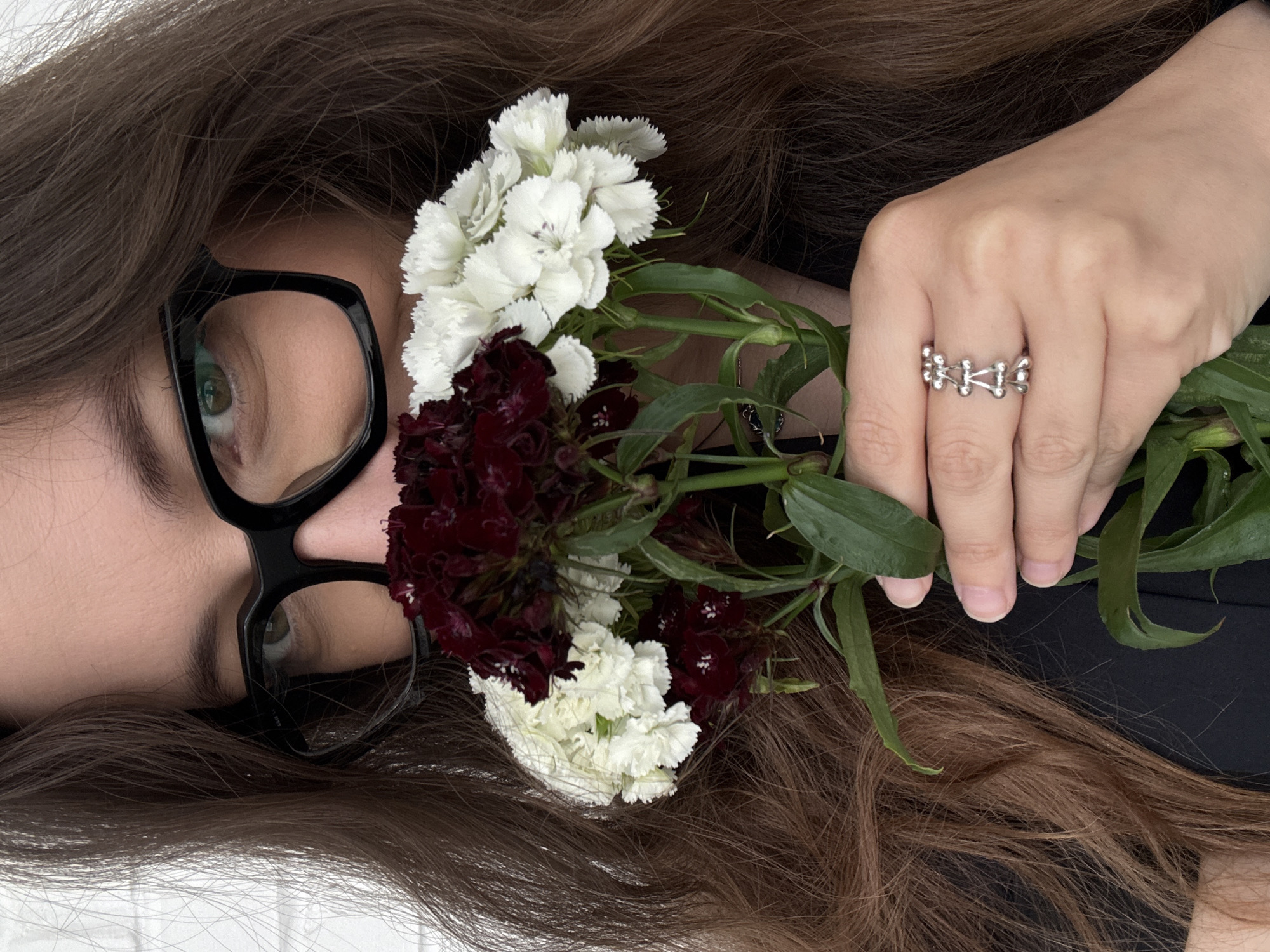 A close-up, medium shot shows a woman with long, brown hair wearing black-rimmed glasses, holding a bouquet of white and dark red flowers to her face. Her eyes are visible behind the glasses, and her mouth is obscured by the flowers. She is wearing a black top and a silver ring with a geometric design on her right hand. The background is a white brick wall. The scene is outdoors. There is no indication of the city of Chaikovsky, Russia.